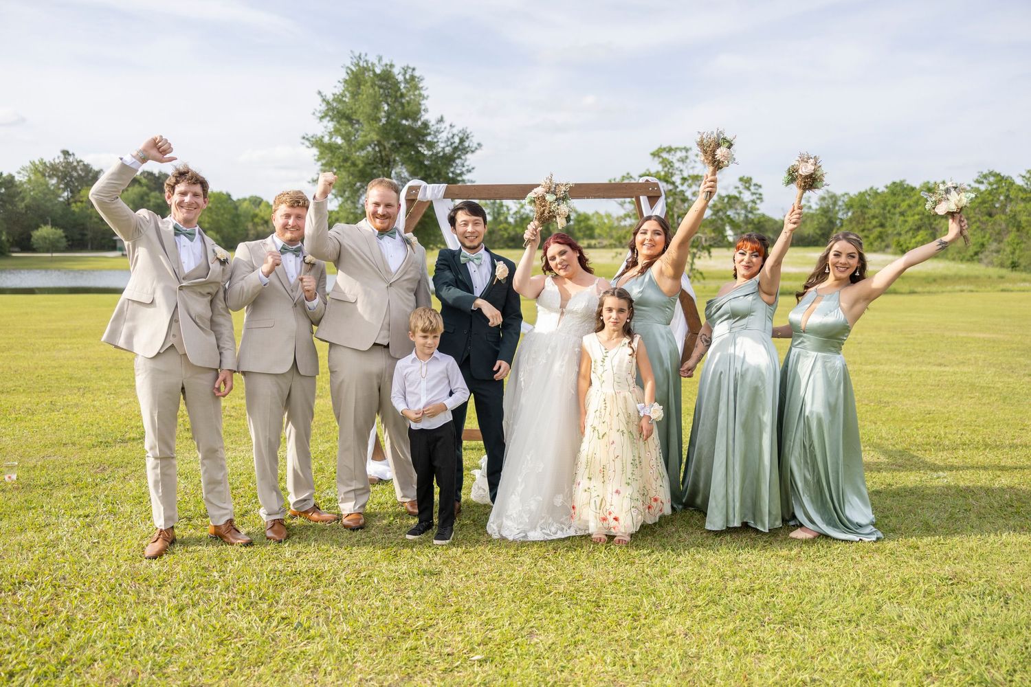 Wedding party in formal attire celebrates joyfully on a green lawn with arms raised against a cloudy sky.
