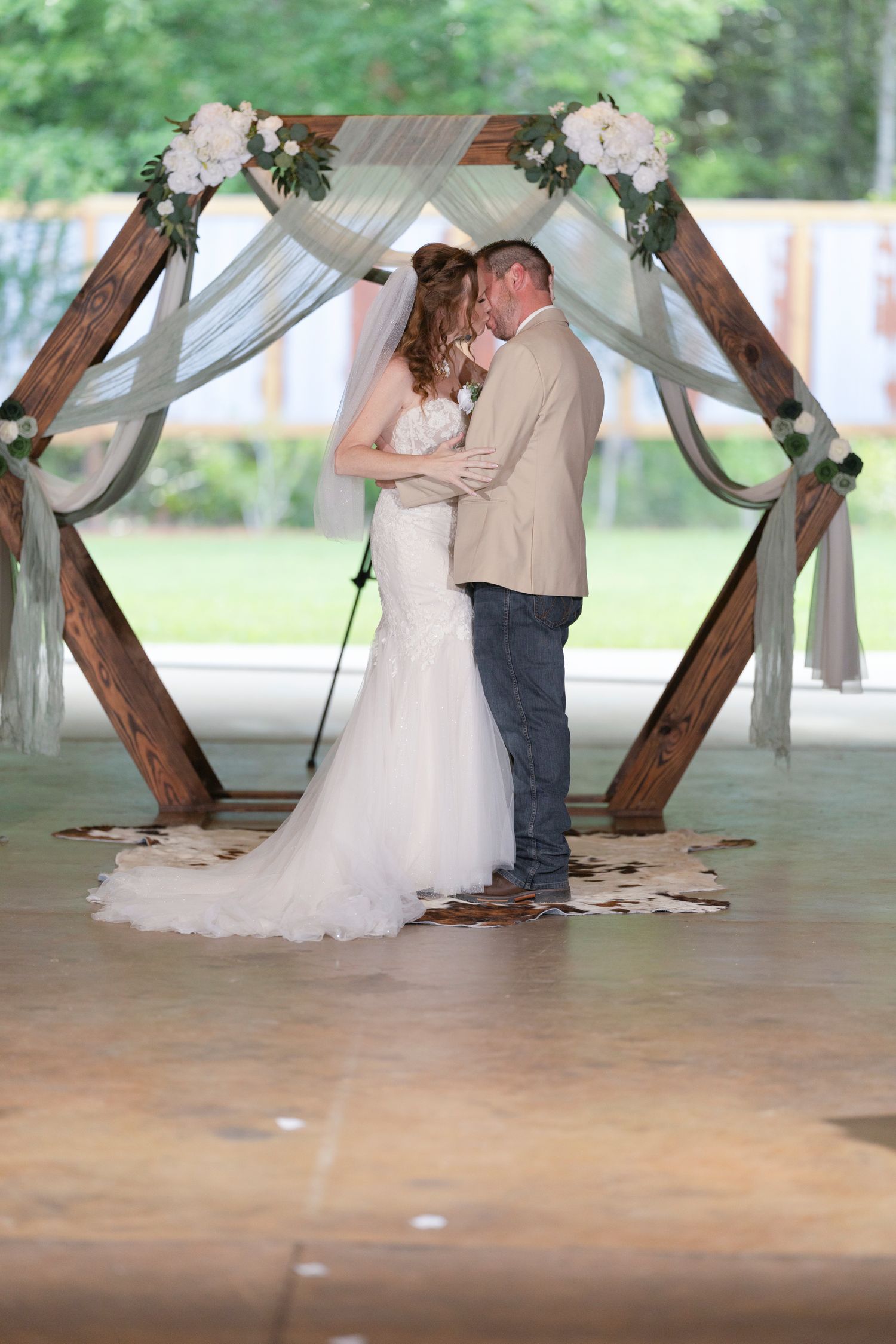 A couple shares a romantic kiss under a rustic wooden arch decorated with flowers and draped fabric at their wedding ceremony.