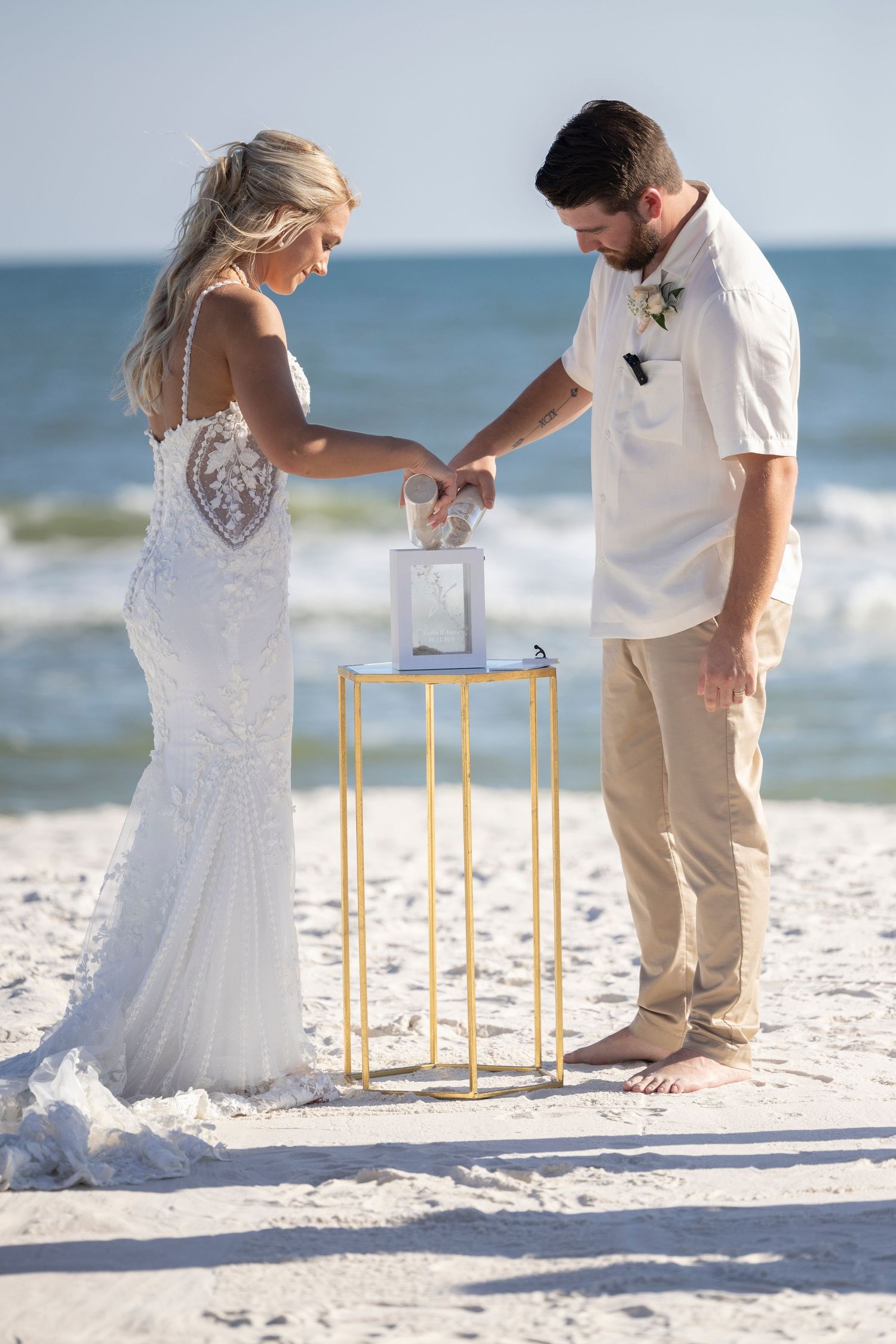 A couple performs a sand ceremony ritual during their beach wedding on a sunny day by the ocean.