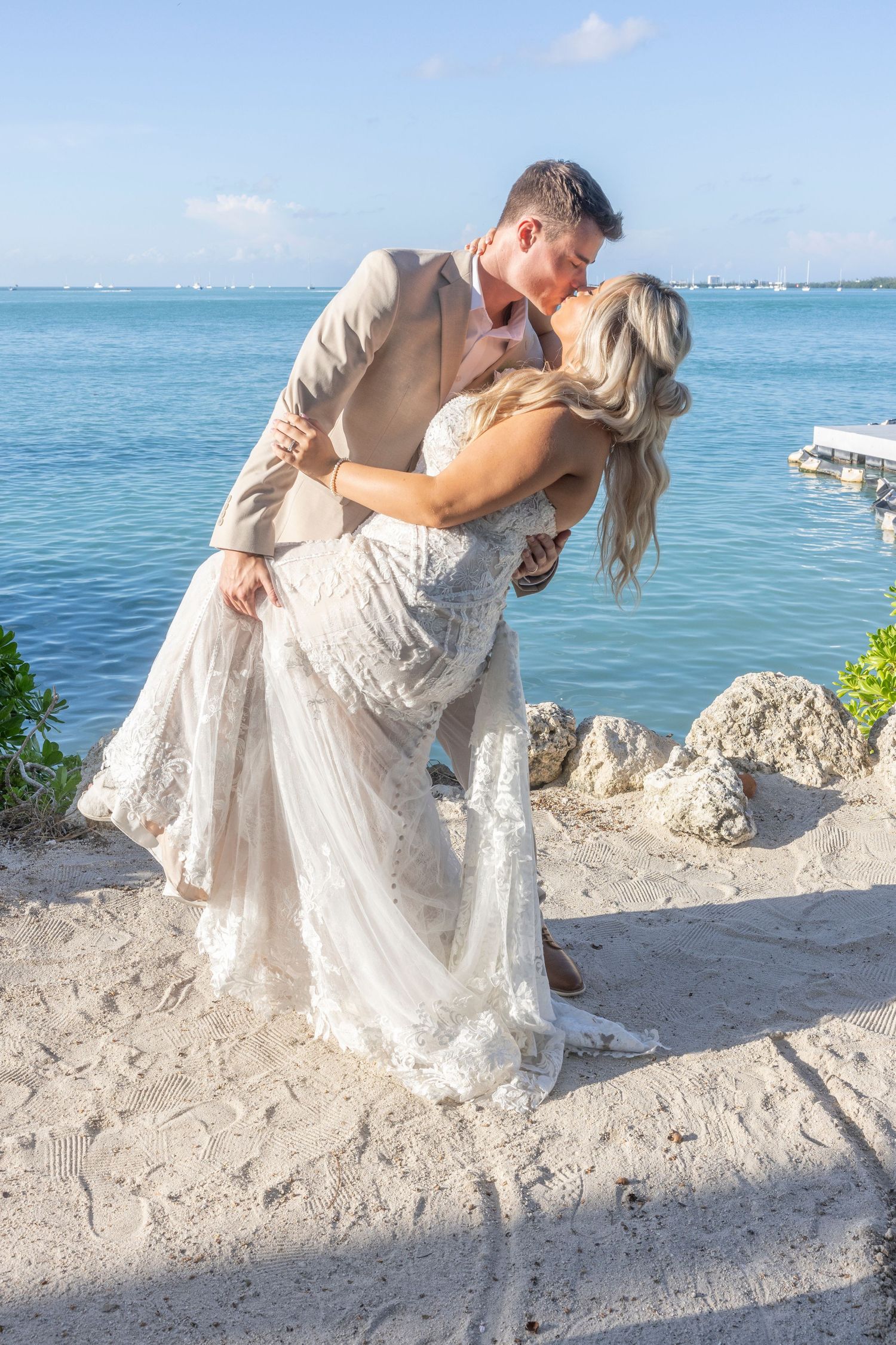A romantic couple shares a tender dip kiss moment on a scenic beach with turquoise waters in the background.
