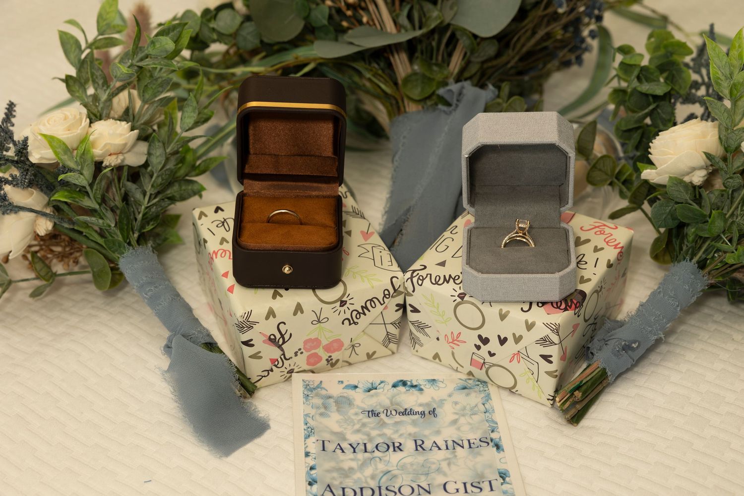 Elegant wedding invitation suite with white roses, greenery, and vintage wooden ring boxes on a cream-colored surface.