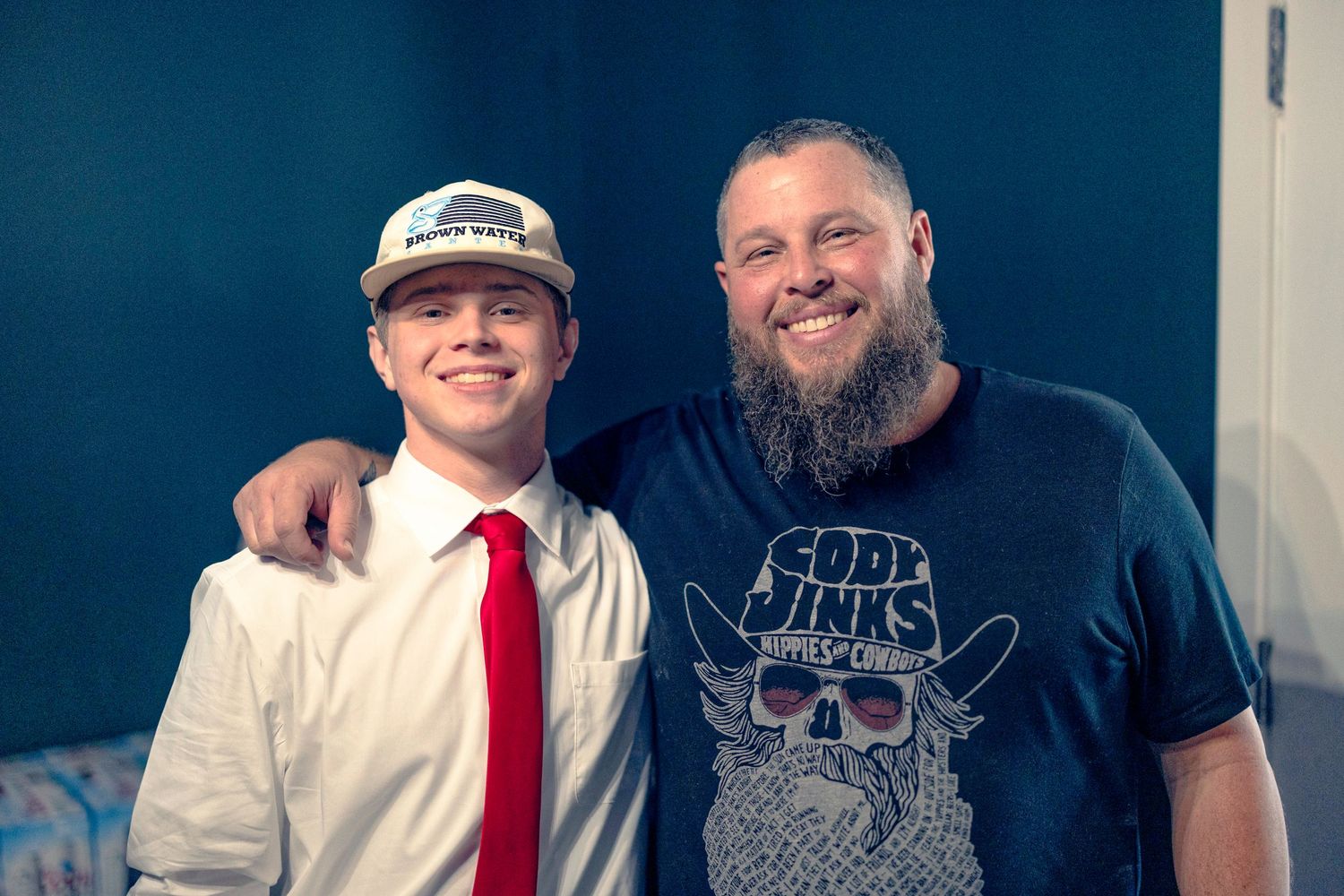 Two people pose together at what appears to be a concert or event venue in a casual setting.
