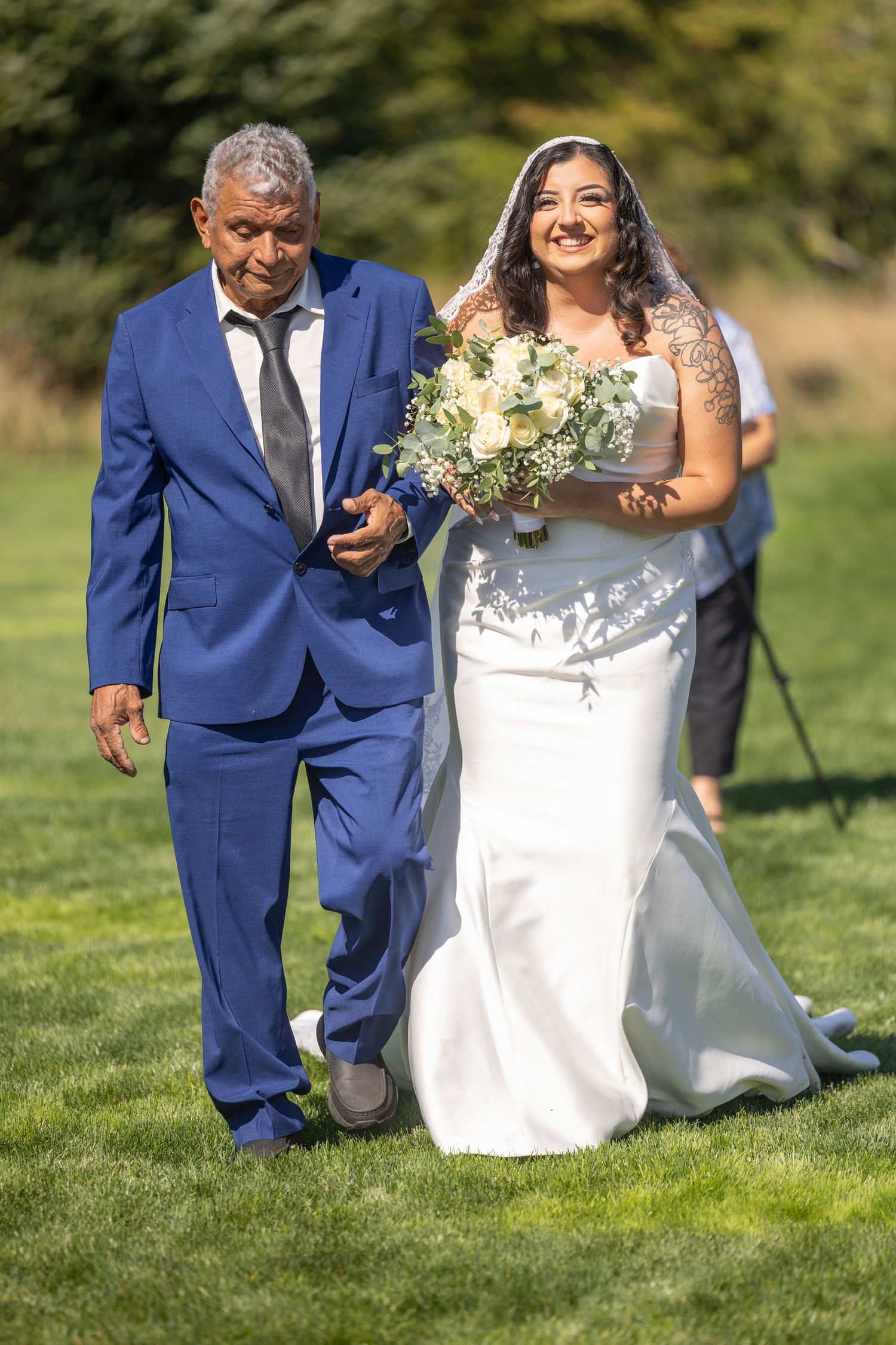 A bride in a white gown walks arm in arm with a companion in a blue suit on a sunny lawn during a wedding ceremony.