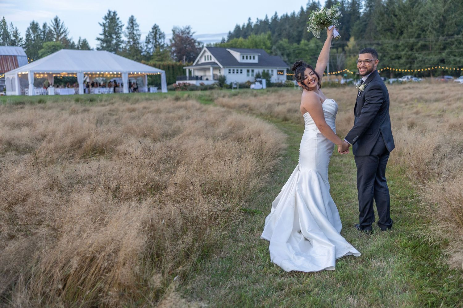 A couple in wedding attire walks through a scenic field of tall grass with evergreen trees in the background.