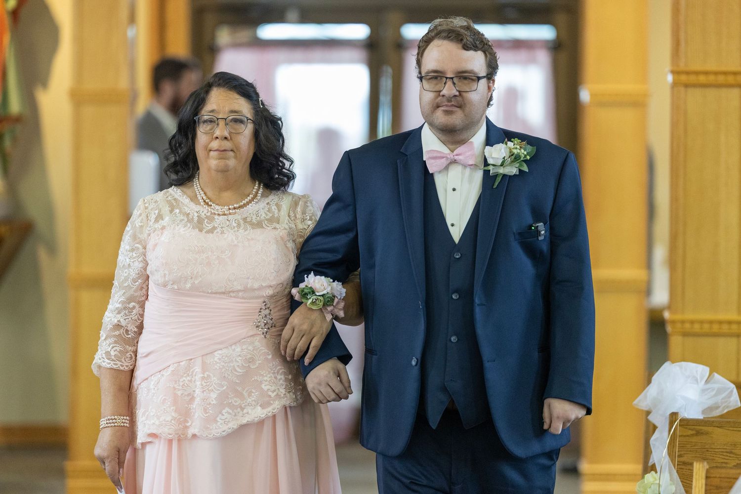 Two people walking arm in arm down a hallway, one wearing a pink lace dress and one in a navy suit.