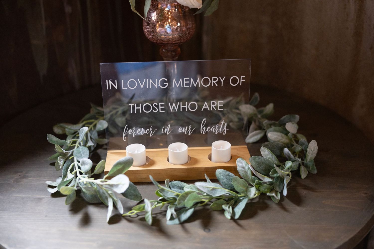 A rustic wooden box with candles and eucalyptus leaves arranged on a dark surface creates a memorial display.