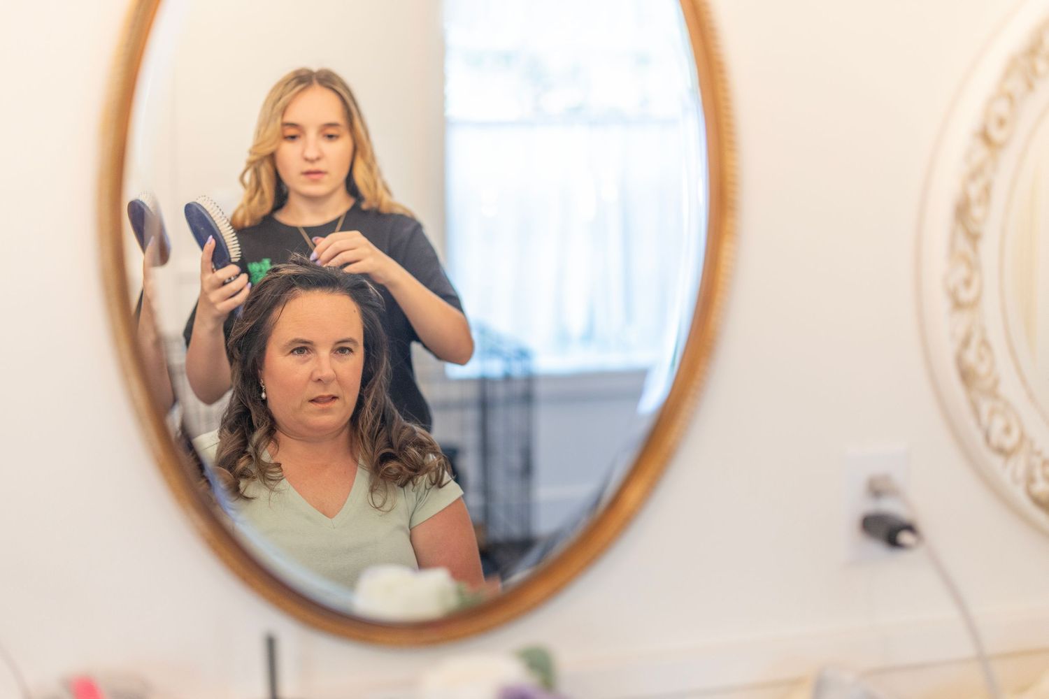Young woman styling curly hair of seated woman, reflected in oval bathroom mirror.