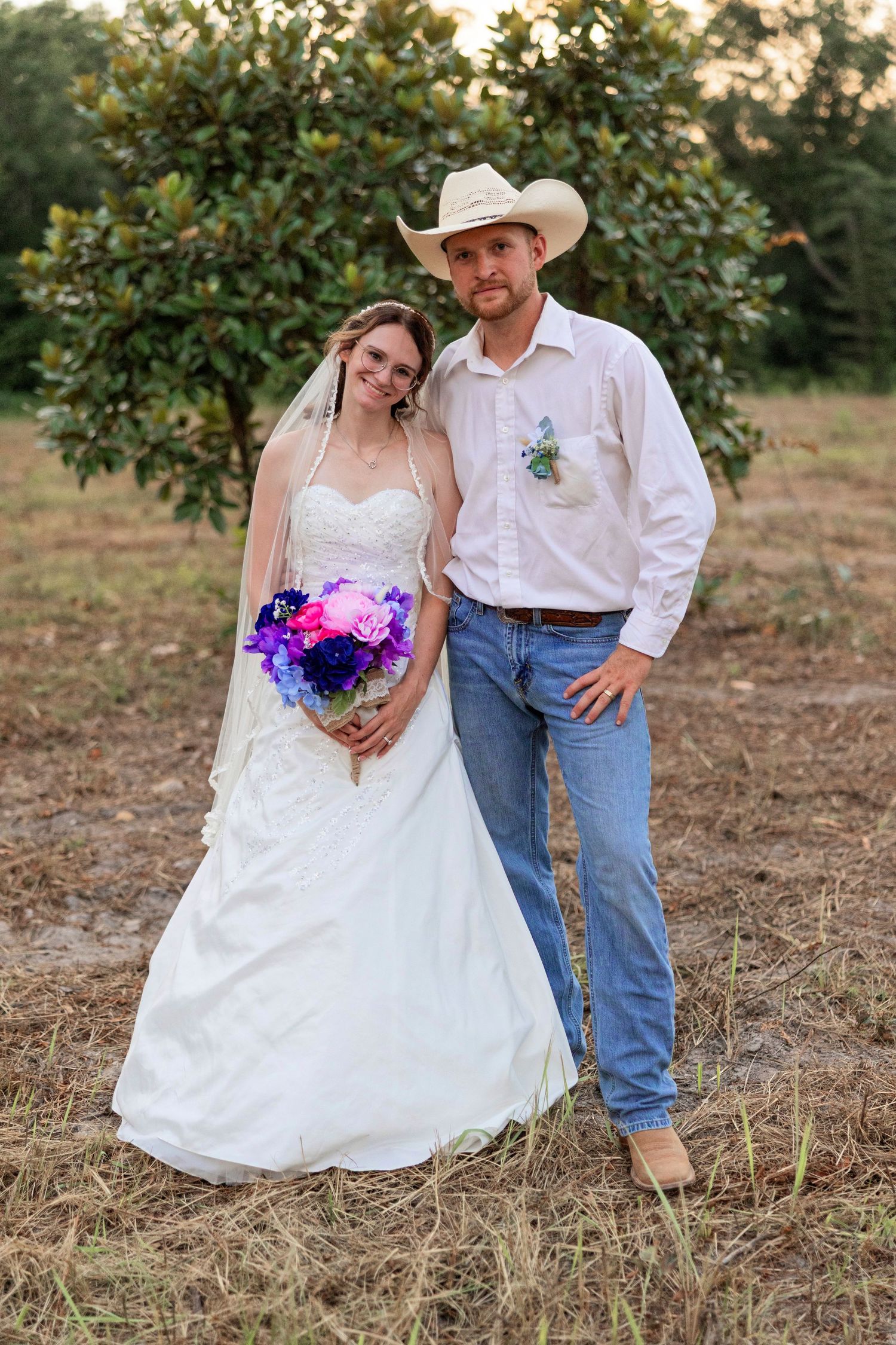 Bride in white gown with purple bouquet and groom in cowboy hat and jeans pose outdoors at rustic wedding.