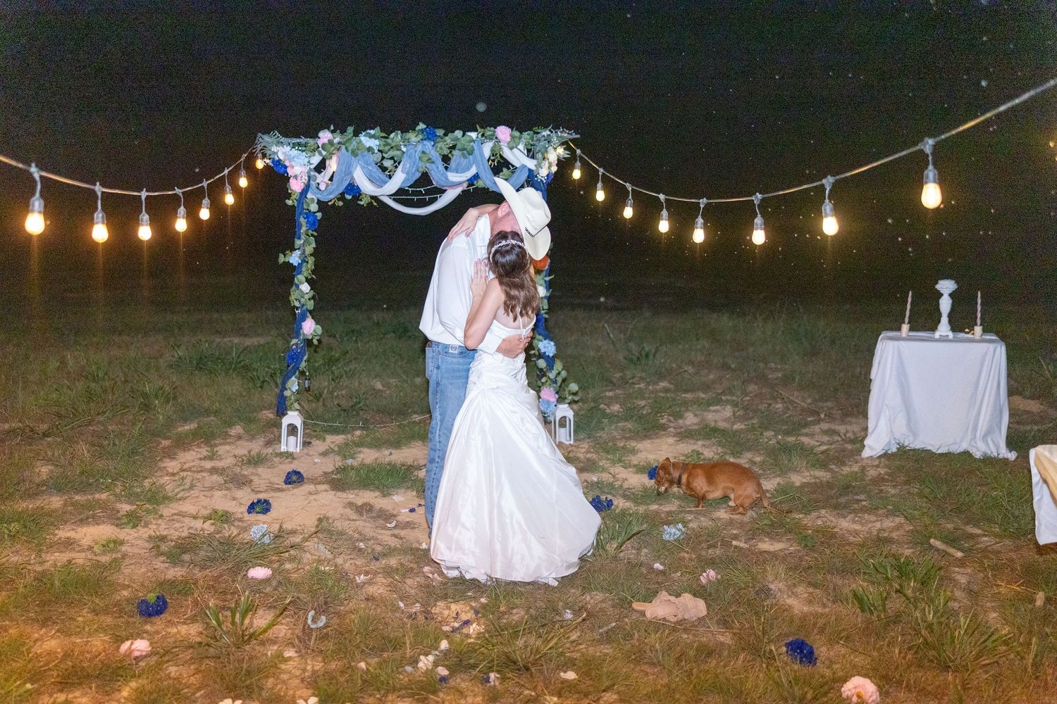 Couple sharing first kiss under floral arch with string lights at outdoor evening wedding ceremony.