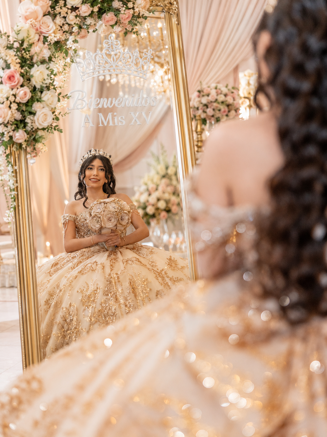 Young woman in gold quinceañera gown and tiara poses before ornate mirror with floral decor.