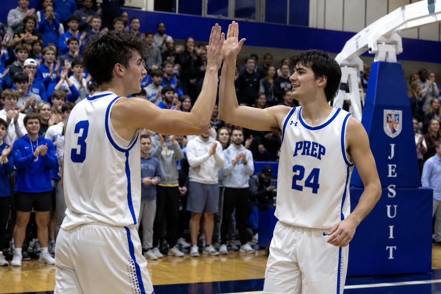 Two Jesuit Prep basketball players in white uniforms high-five on court as crowd cheers.
