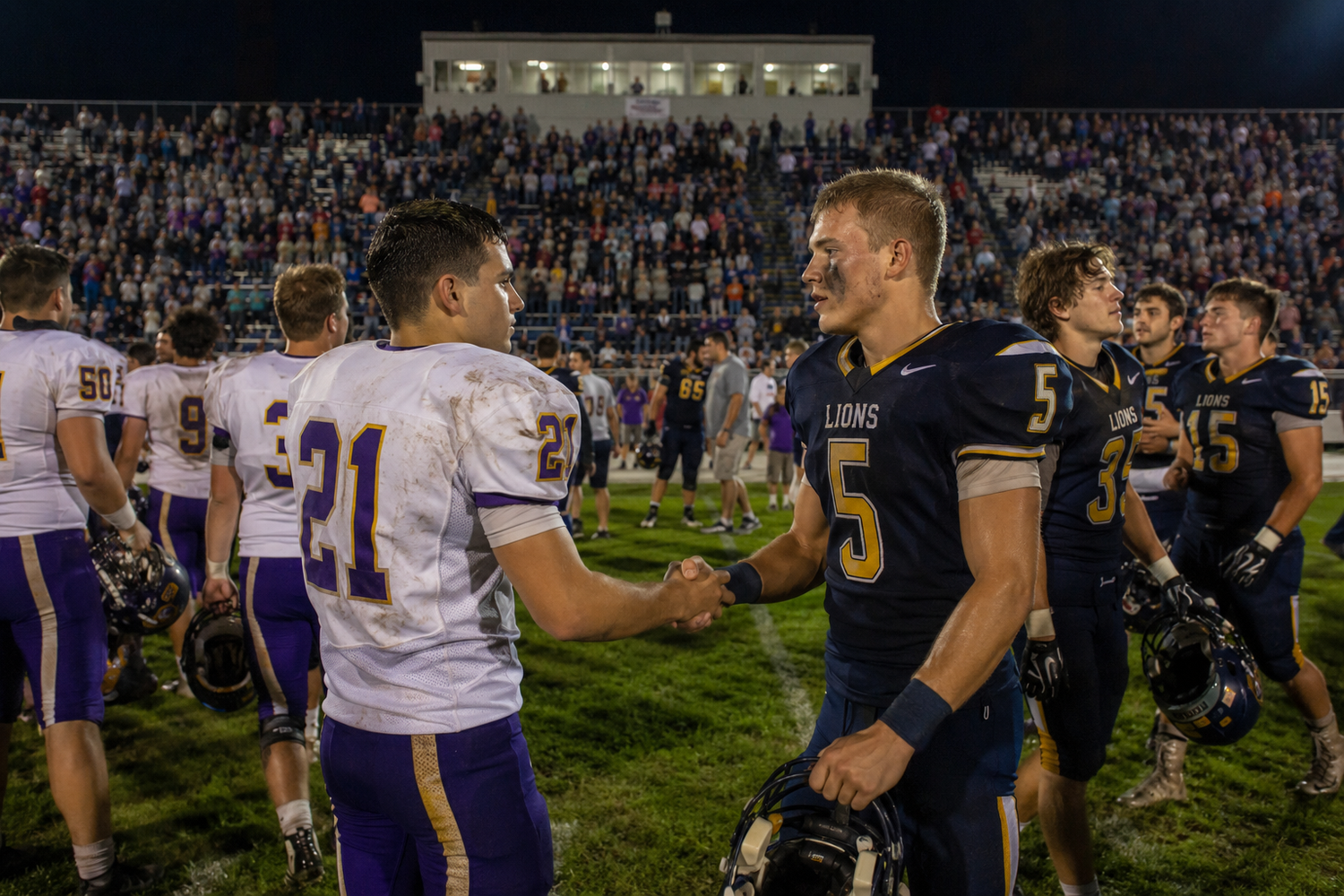 Football players from opposing teams shake hands after game before packed stadium crowd.