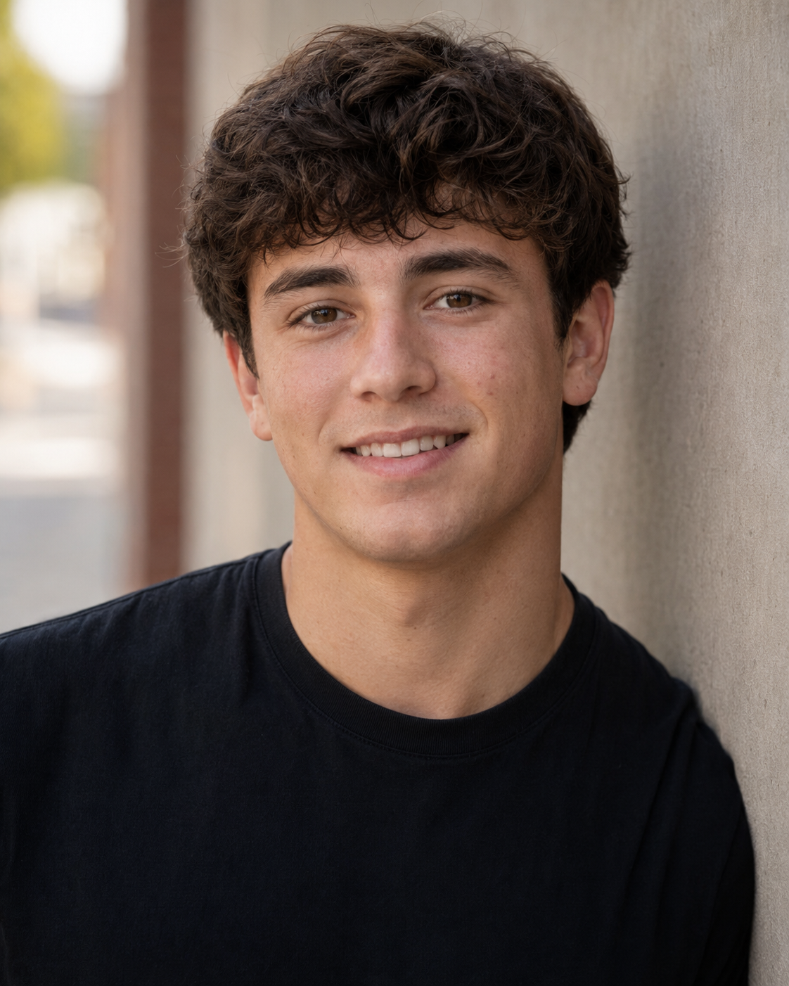 Smiling teenage boy with curly brown hair wearing a black t-shirt in an outdoor portrait photo.