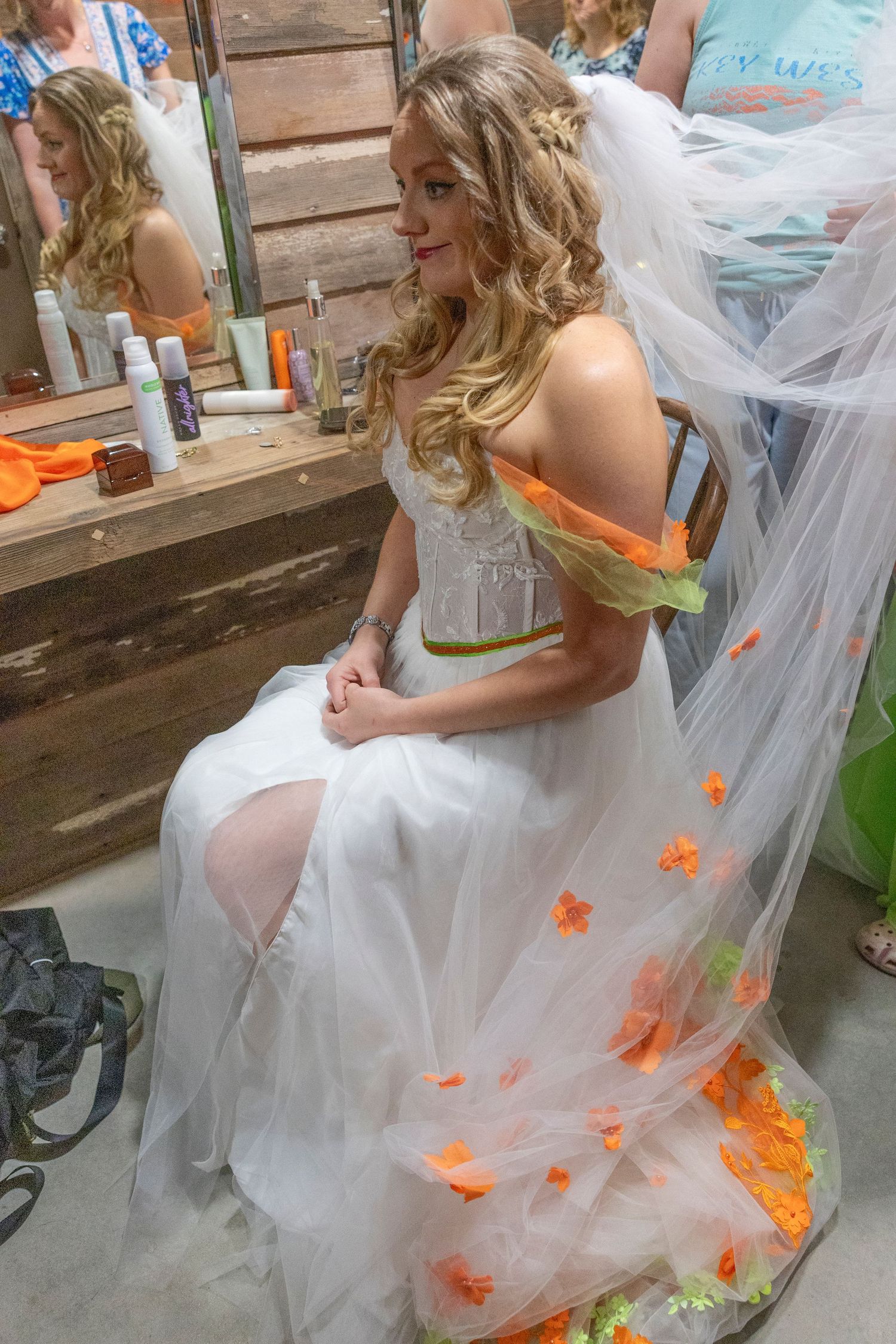 Bride in white tulle gown with orange floral accents sits before a mirror in a rustic dressing room.