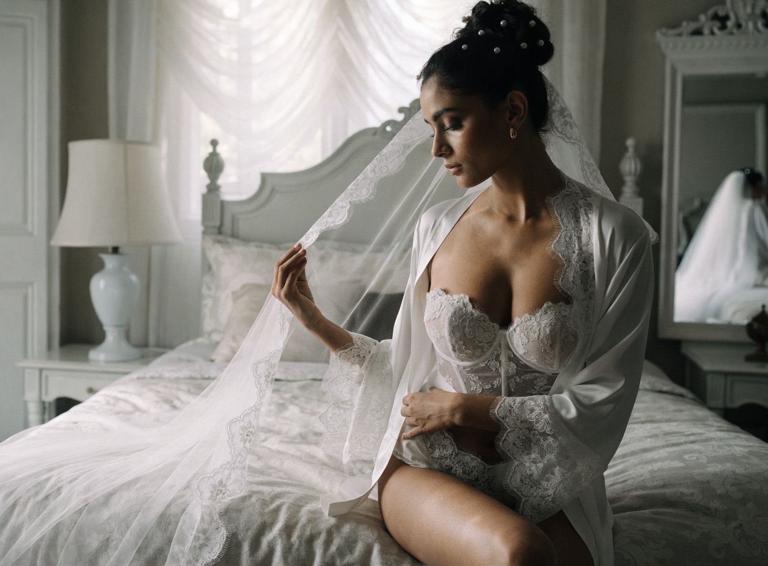 Bride in lace corset and sheer robe holding wedding veil while sitting on bed in elegant bedroom.