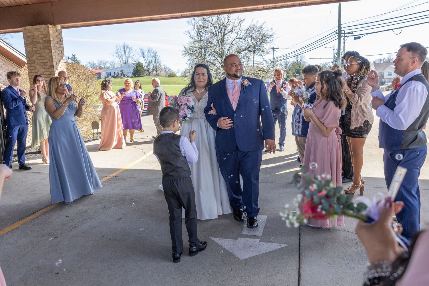 Newlywed couple walks through cheering guests at outdoor wedding ceremony, bride in white gown, groom in blue suit.