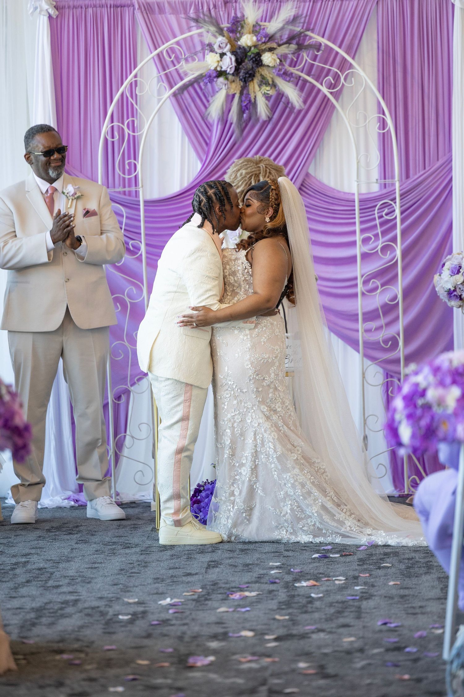 Newlywed couple shares first kiss at altar with purple draped arch backdrop during elegant wedding ceremony.