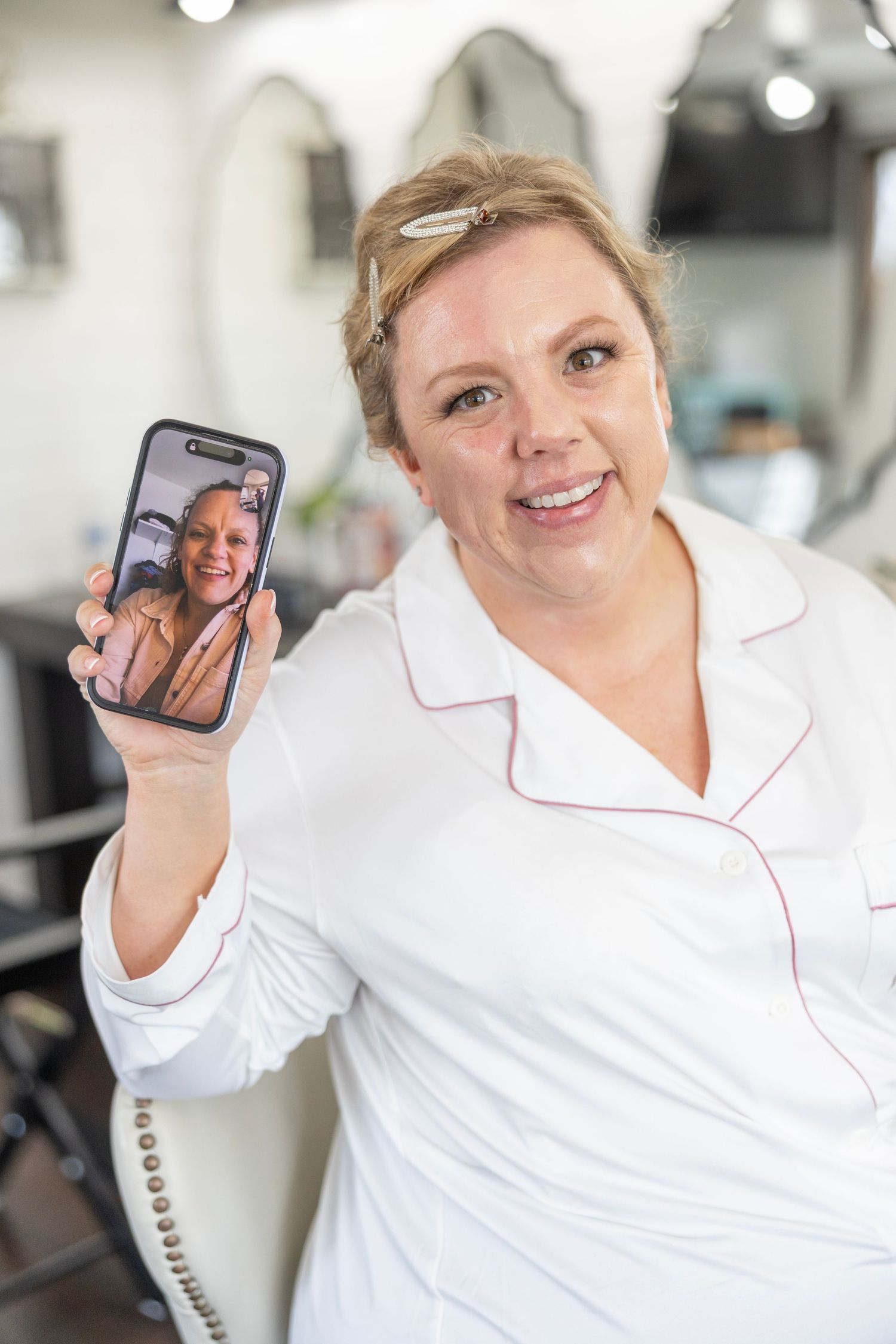 Woman in white salon coat smiling while holding up smartphone showing FaceTime call in hair salon.