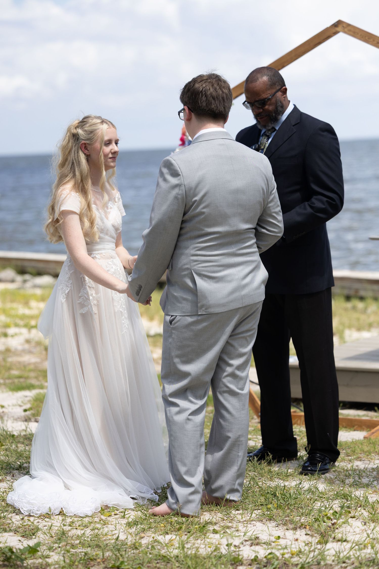 A wedding ceremony takes place on a scenic coastal shoreline with an officiant and couple in formal attire.