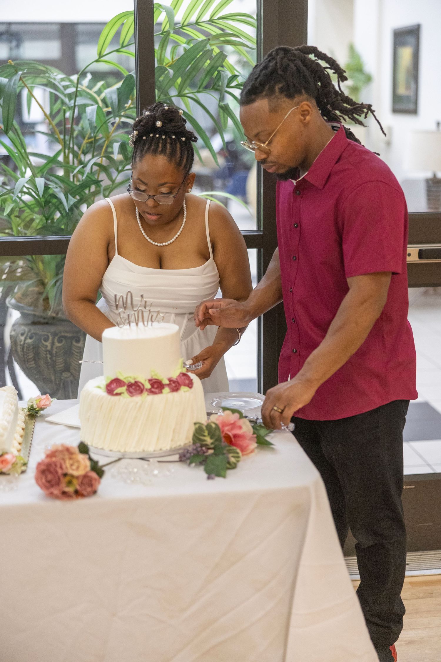 A couple cutting their white wedding cake decorated with red flowers at an intimate indoor celebration.