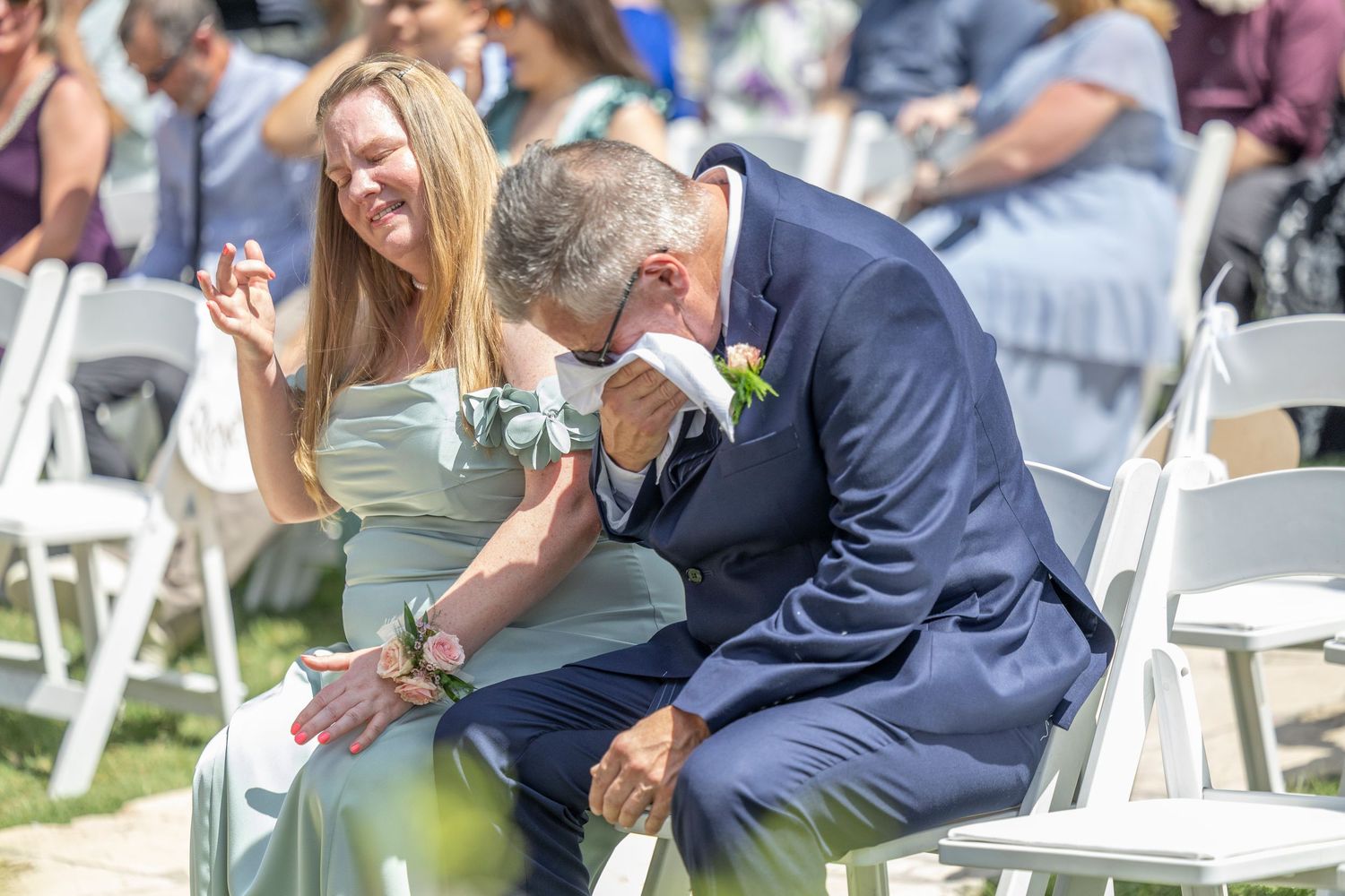 Wedding guests share laughter during an outdoor ceremony with white chairs on a sunny day.