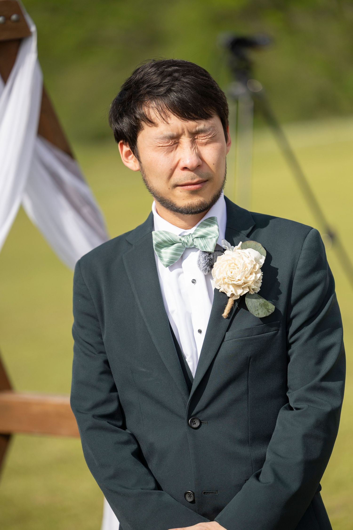 A groom in a dark suit and white bow tie displays a range of emotional expressions during an outdoor wedding ceremony.