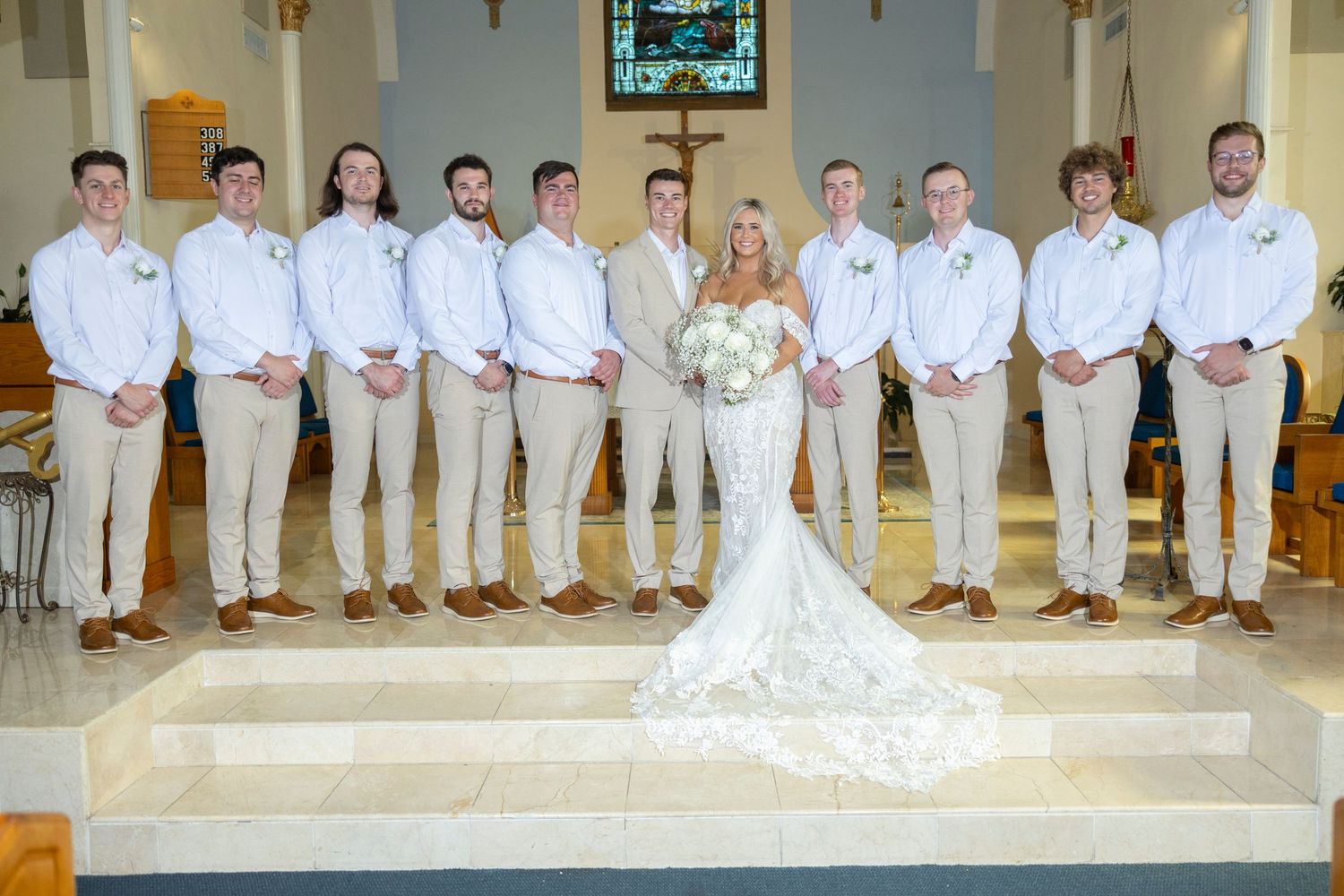 Wedding party members in white and khaki attire pose together on church steps for formal group photo.
