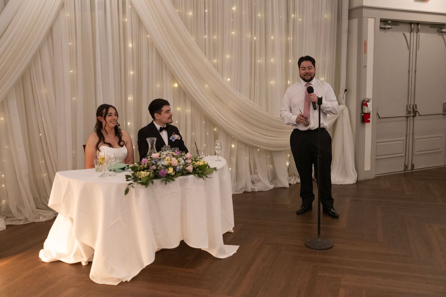 A groom gives a wedding toast at a microphone while the bride and groom sit at a decorated head table with white draping.