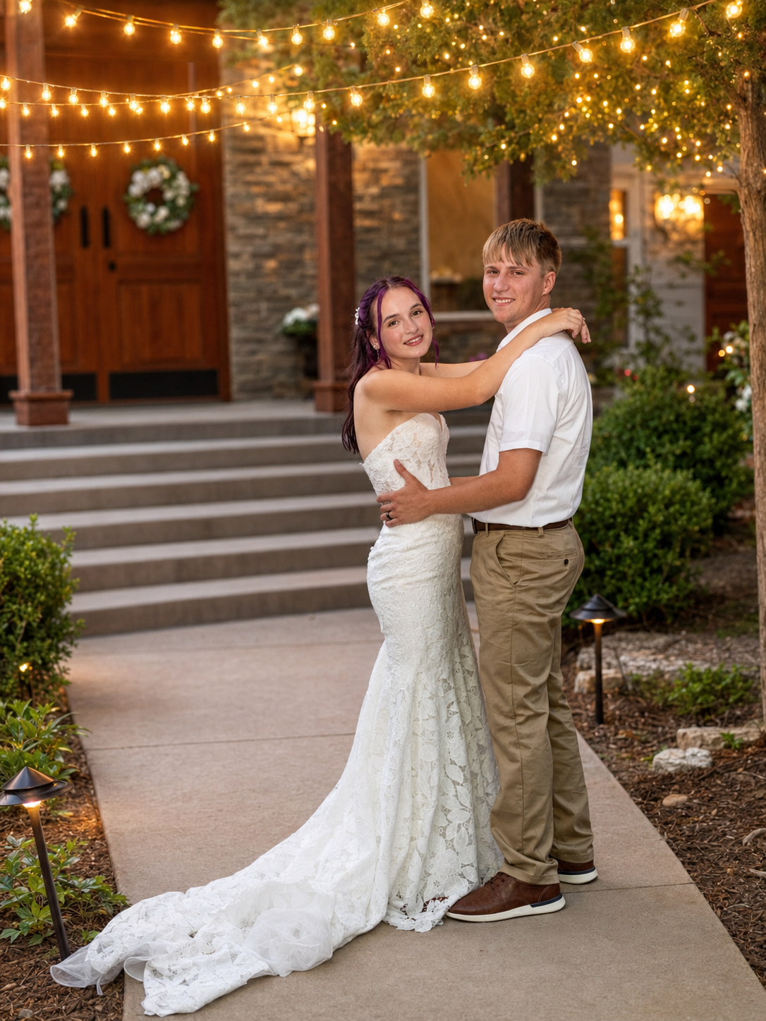 Bride in white lace gown and groom in khakis pose under string lights outside stone venue at dusk.