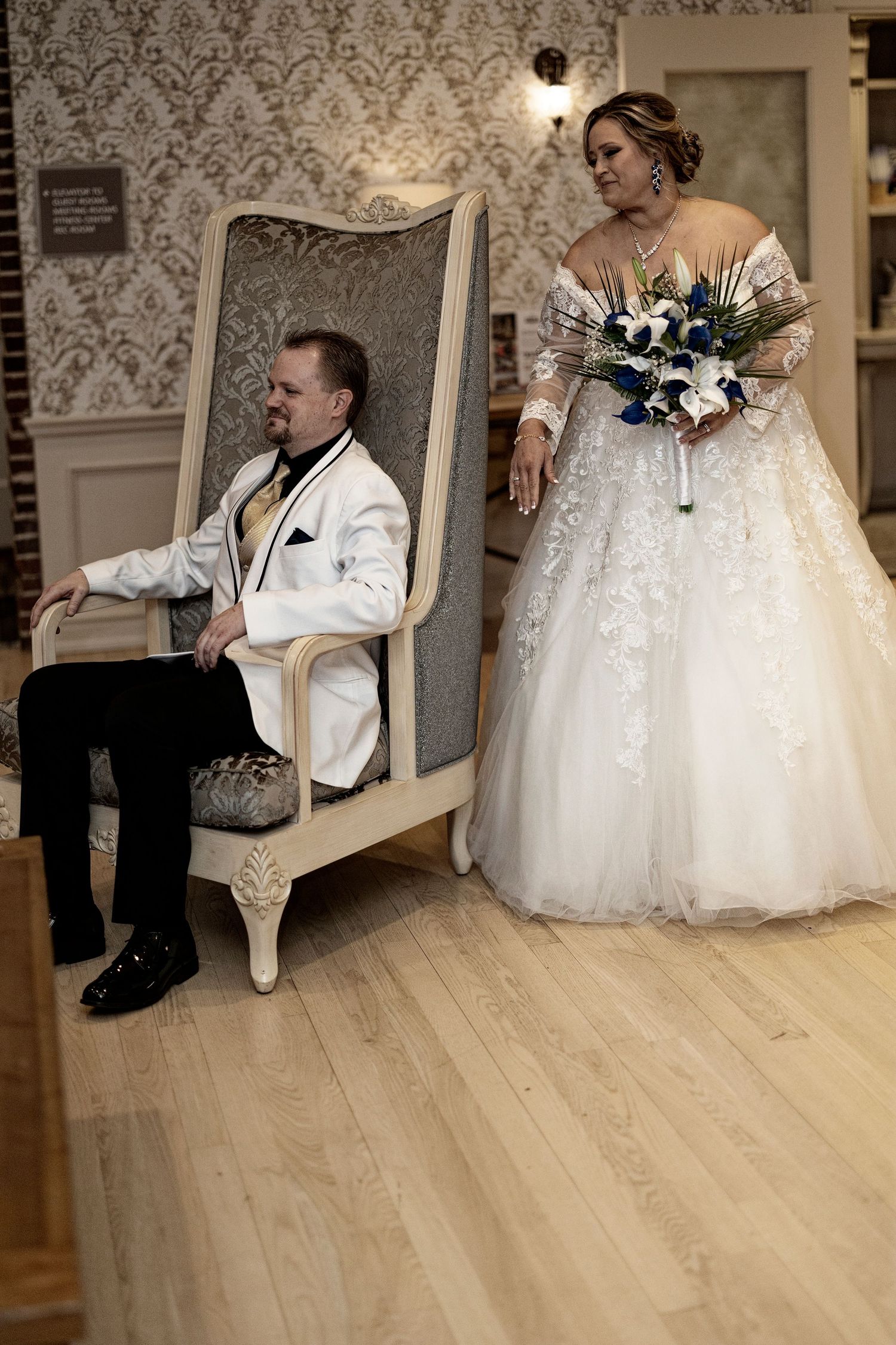 Bride in white lace ball gown holding blue bouquet stands beside groom in white tuxedo seated in ornate chair.