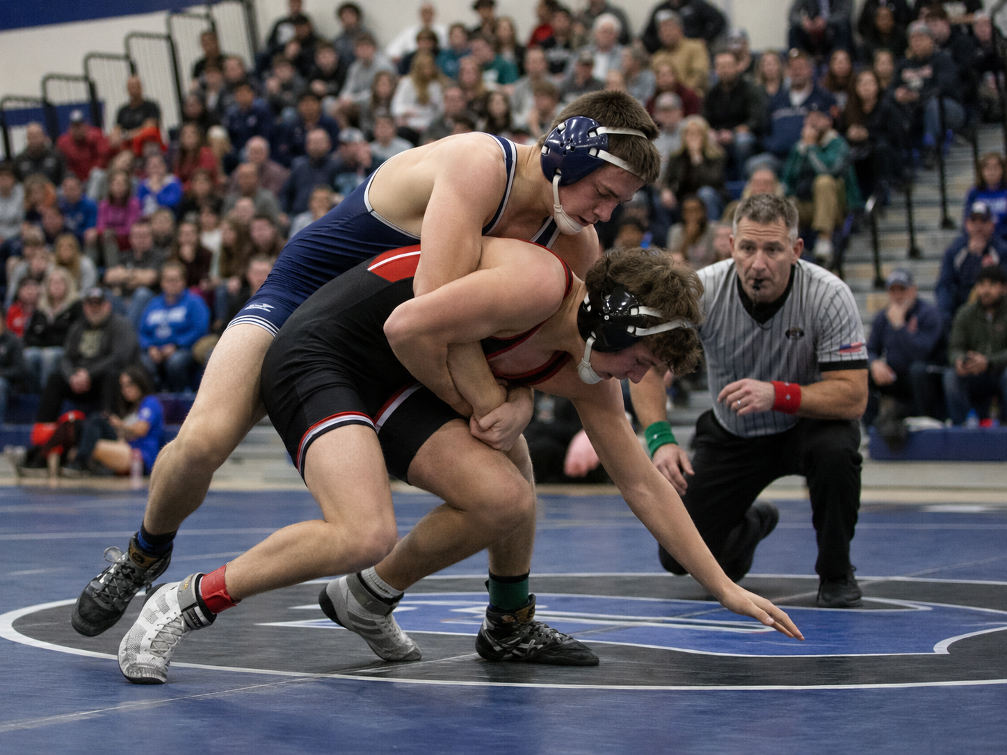Two wrestlers grapple on the mat as a referee watches closely during a high school wrestling match.