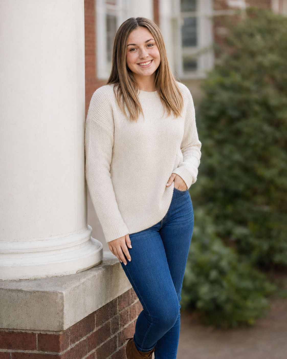 Smiling young woman in a cream sweater and blue jeans leaning against a white column outdoors.