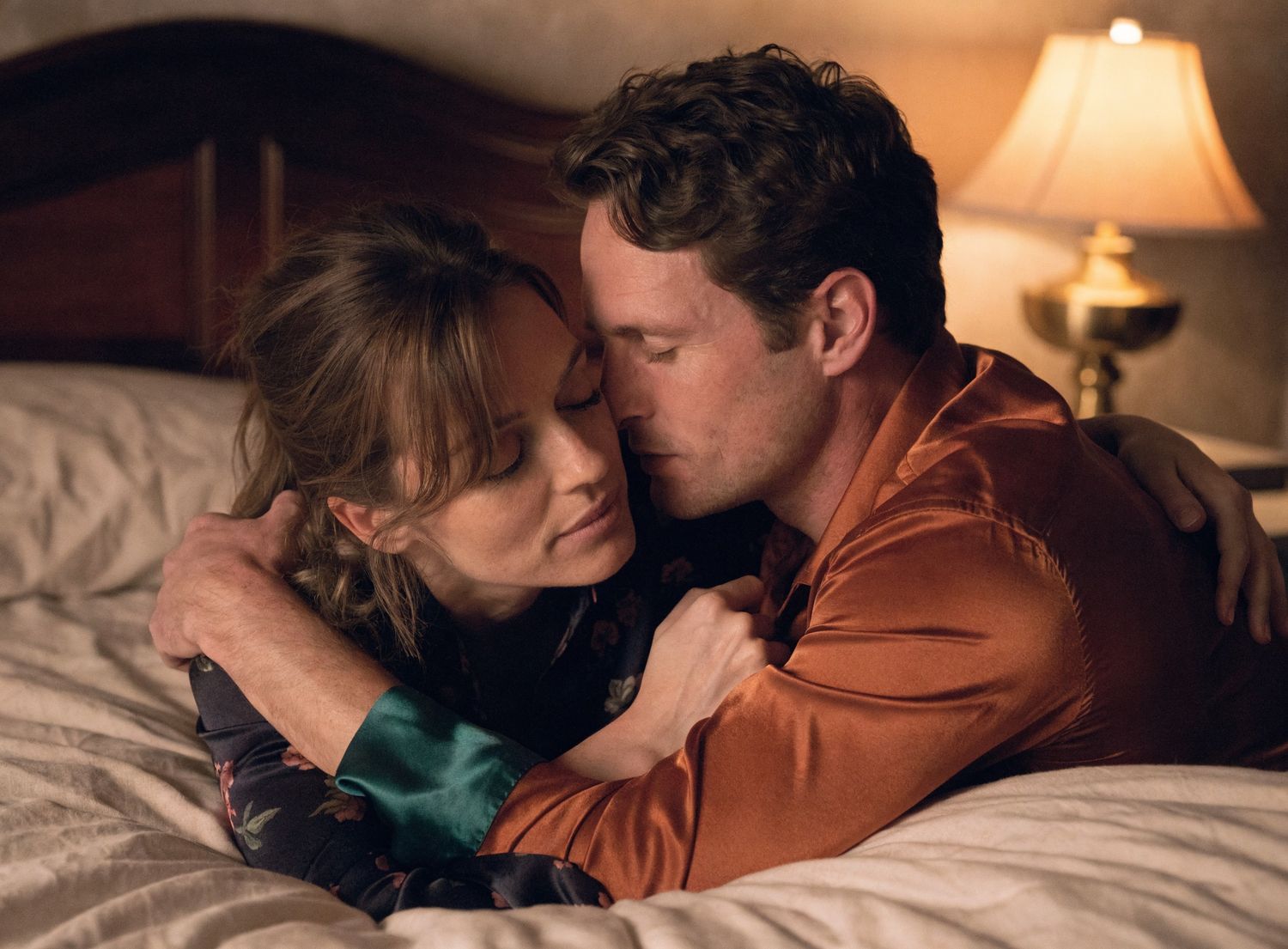 A couple shares an intimate embrace while lying on a bed in a warmly lit room.