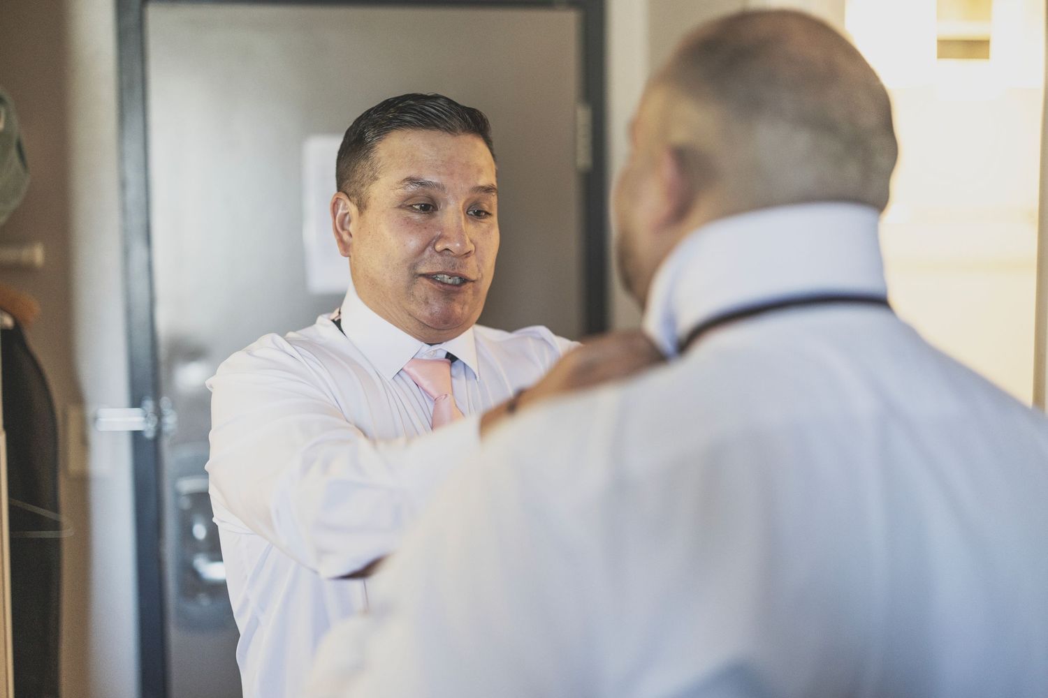 Two men in white dress shirts adjusting ties, preparing for a formal event in a well-lit room.