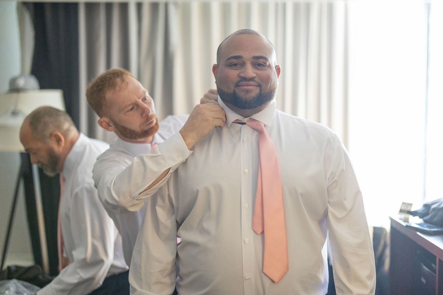 Groomsmen helping each other get ready, adjusting bow tie on wedding day in hotel room.