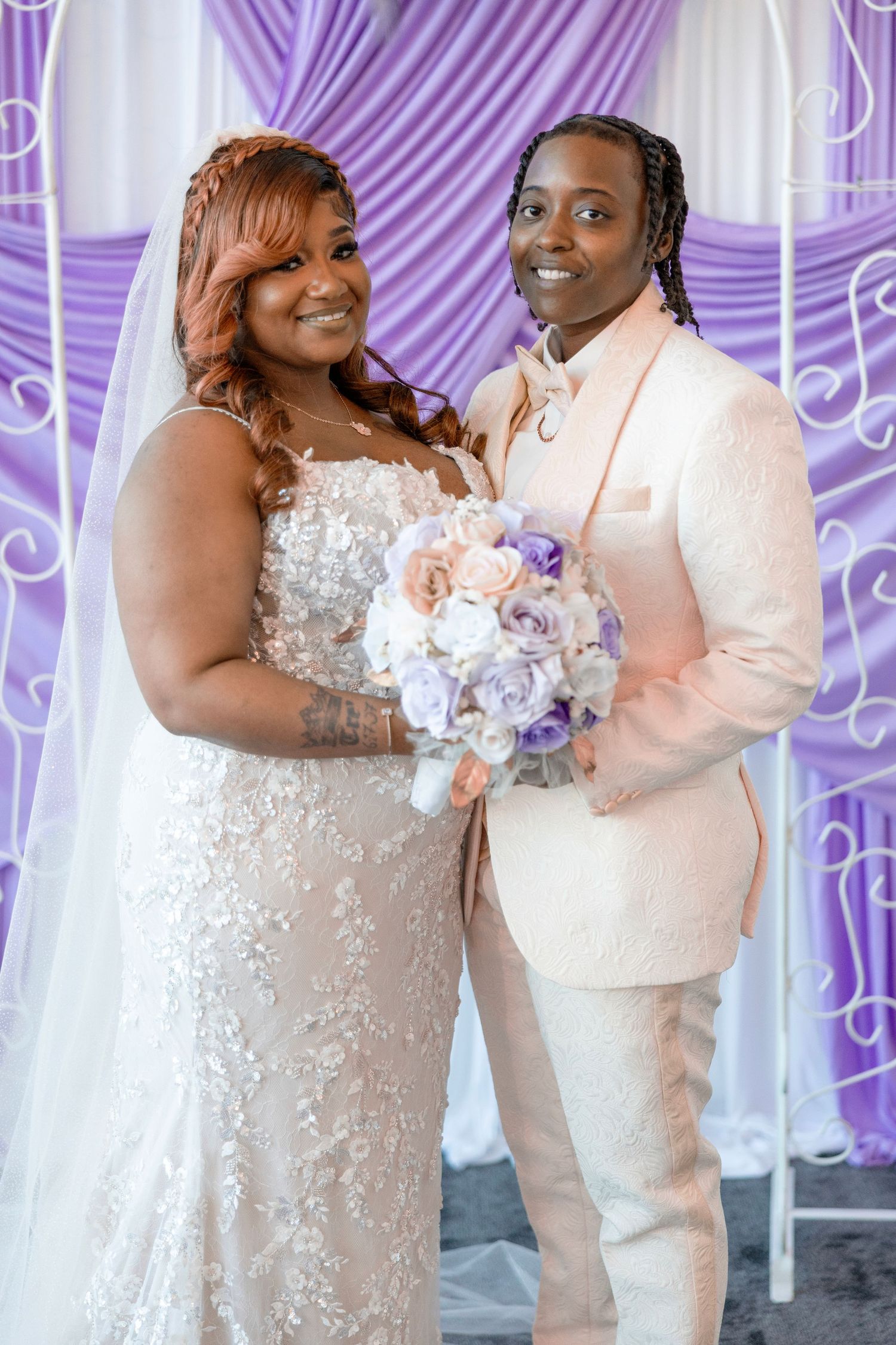 Two brides pose together at their wedding, one in a lace gown and one in a cream suit, holding purple and pink bouquets.