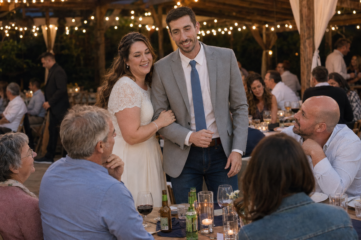 Couple in wedding attire smiling and mingling with guests at an outdoor reception with string lights.