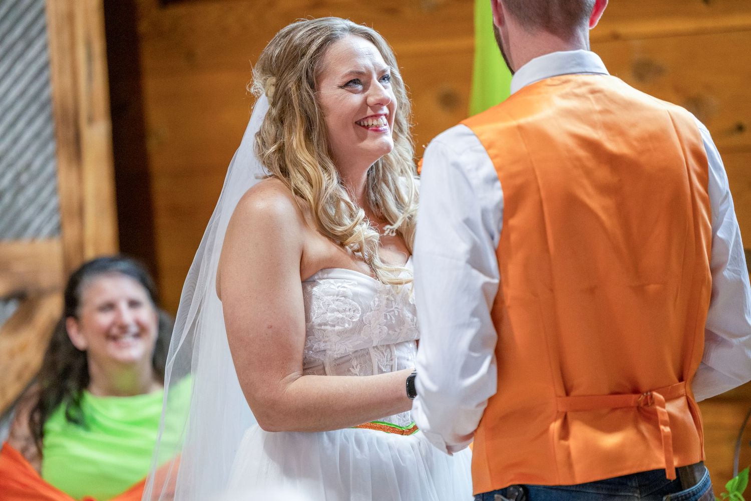 Smiling bride in white lace gown and veil exchanges vows with groom in orange vest at rustic outdoor wedding ceremony.