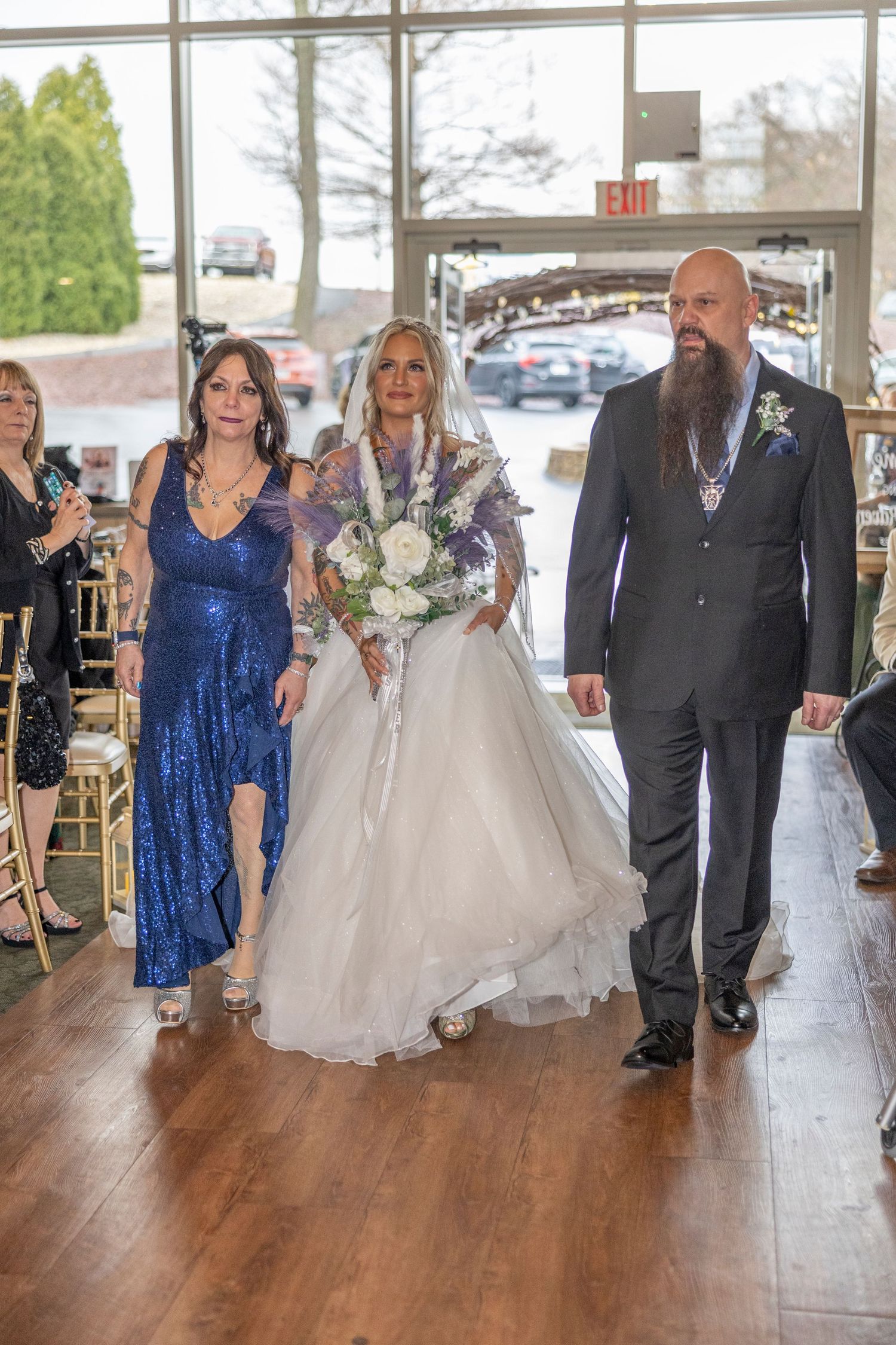 Bridal procession walks down hardwood floor aisle, with attendant in sparkling blue dress and groom in dark suit.
