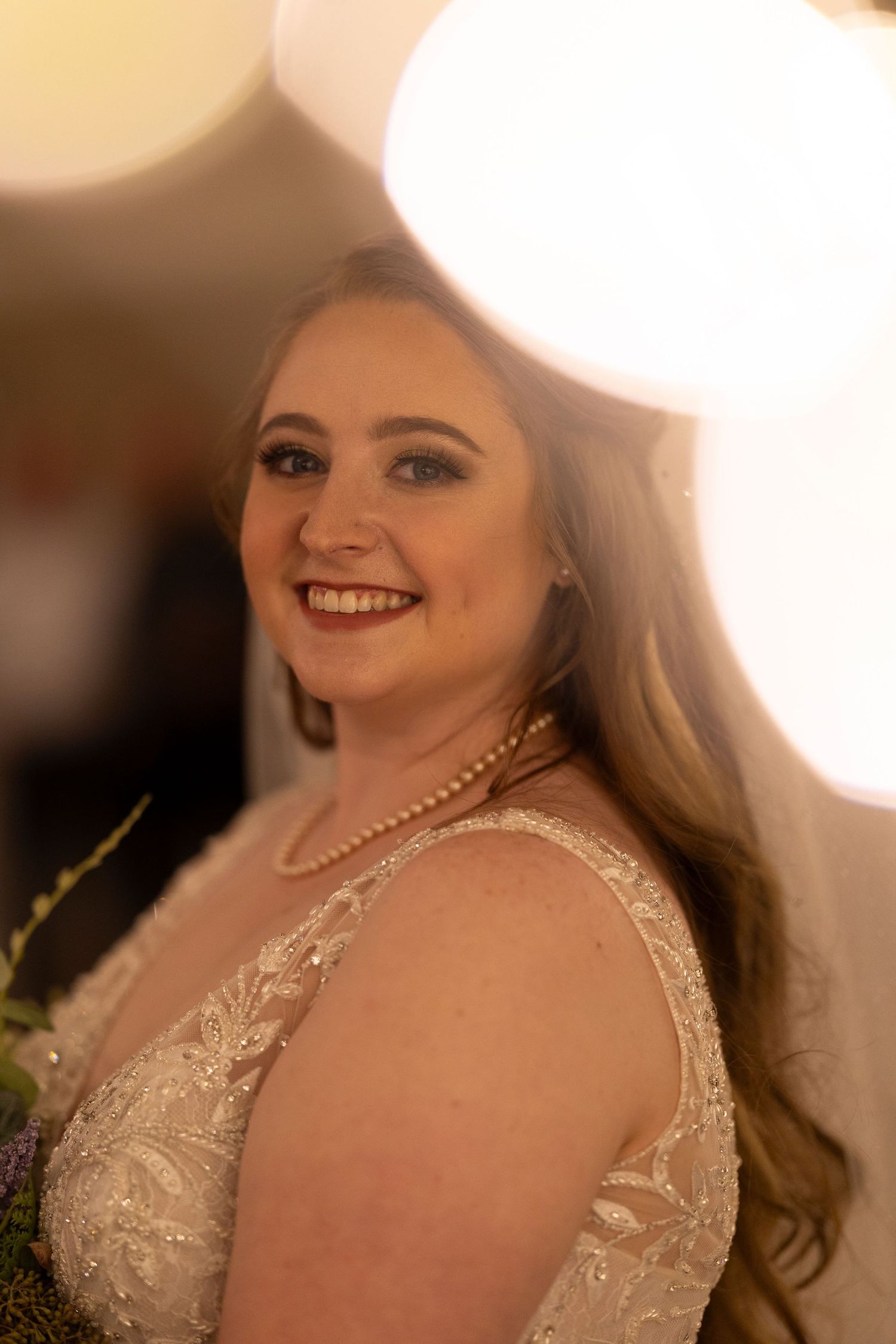 A bride wearing pearls and an embellished wedding gown smiles in soft lighting.