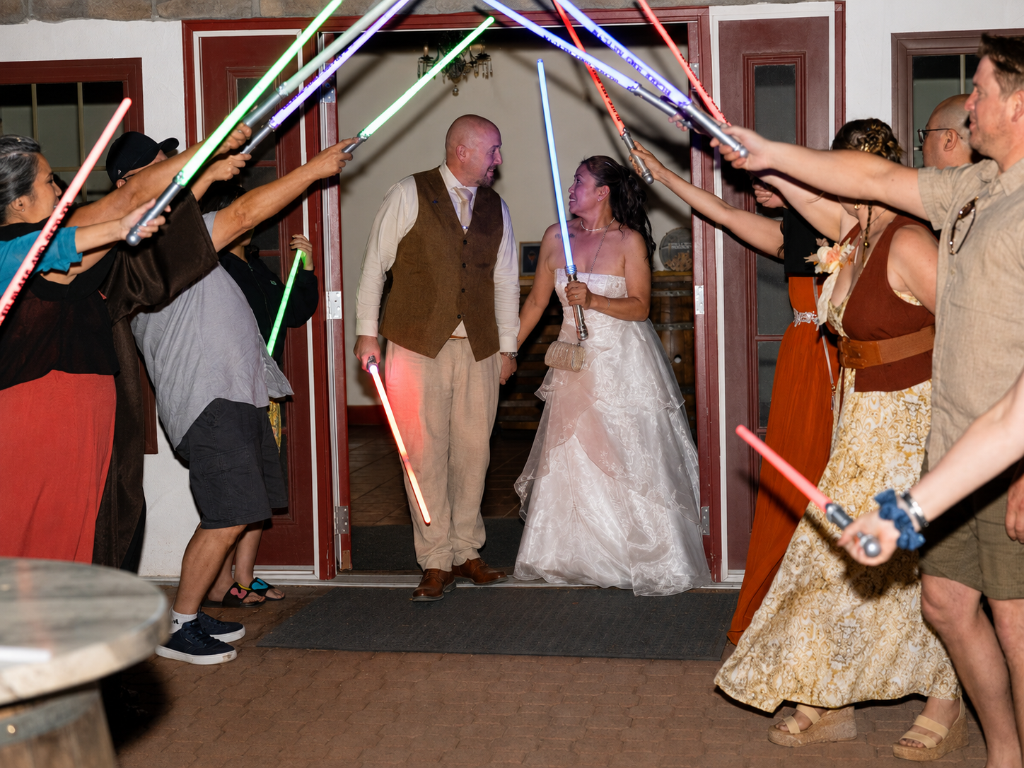 Star Wars themed wedding couple exits under crossed lightsabers held by guests in colorful celebration.