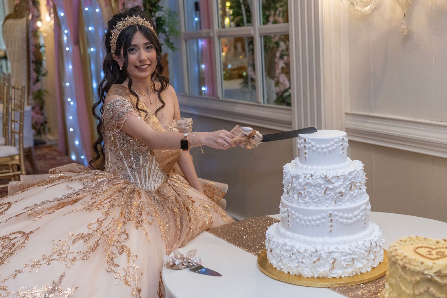 Young woman in gold quinceanera gown cutting a white four-tier cake at an elegant celebration venue.