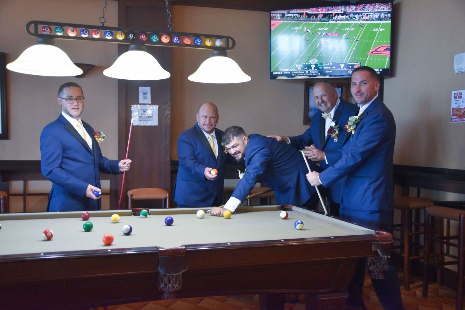 A group in blue suits playing pool at a bar with a TV showing football in the background.