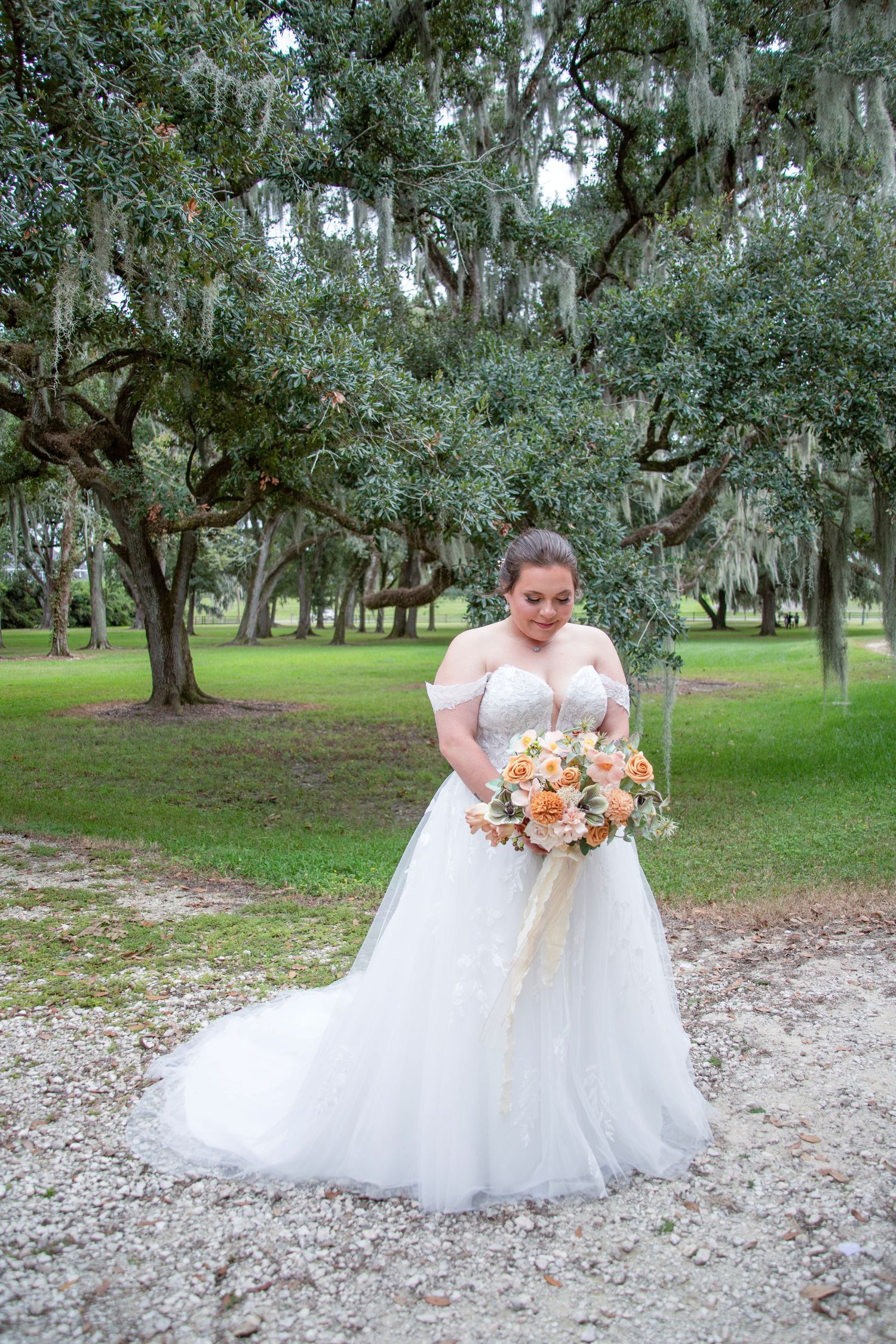 A bride in a flowing white wedding dress poses gracefully under Spanish moss-draped oak trees in a serene outdoor setting.