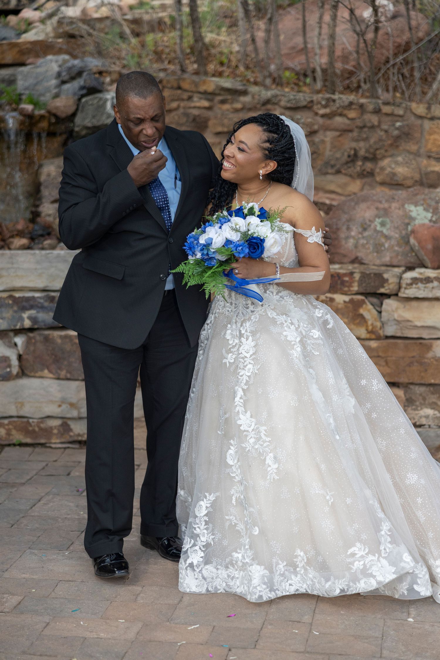 A bride in a white lace ball gown shares a romantic moment with her partner against a stone wall backdrop.