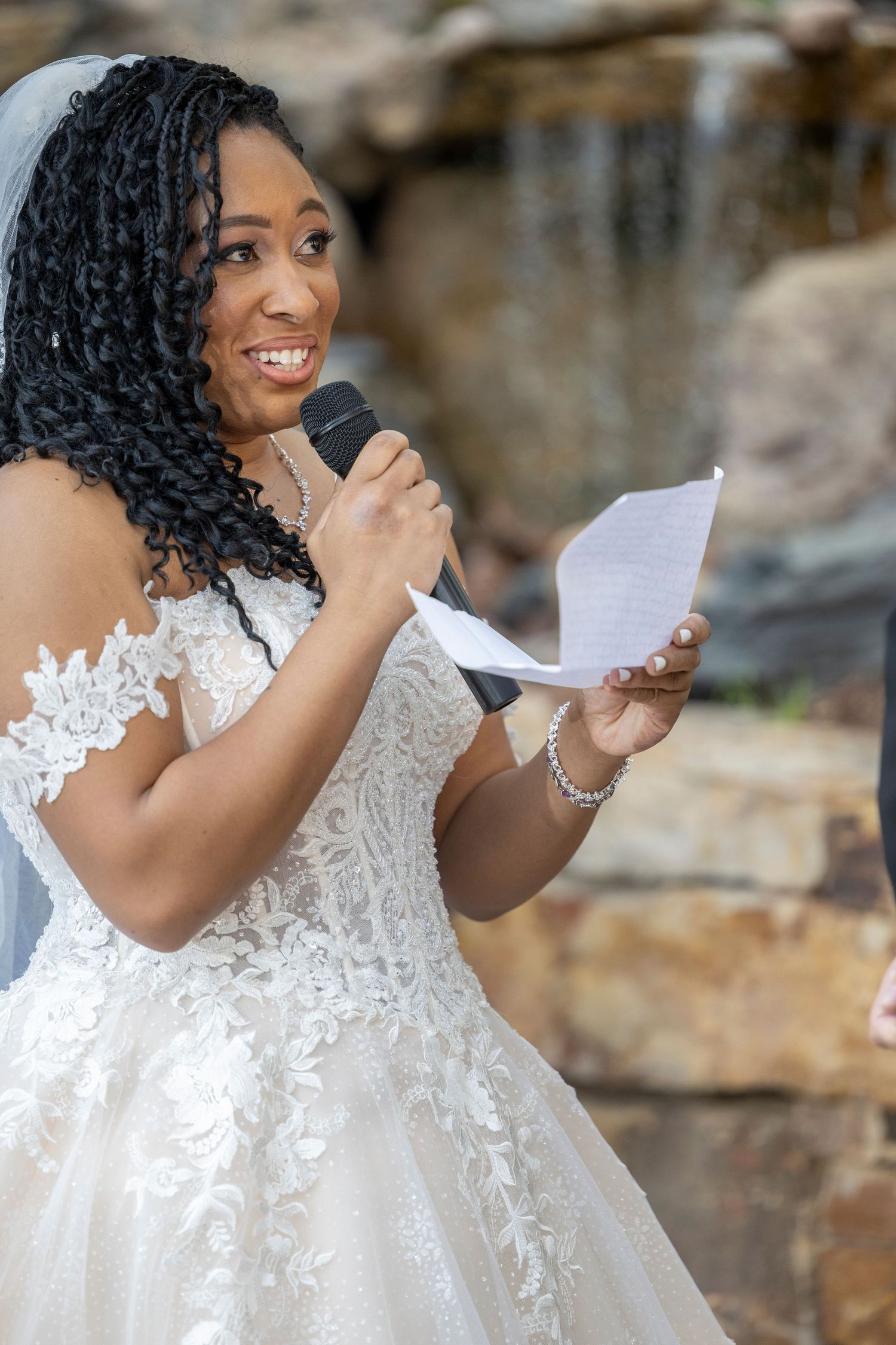 A person in a white lace wedding dress reads a letter while posing against a stone wall background.