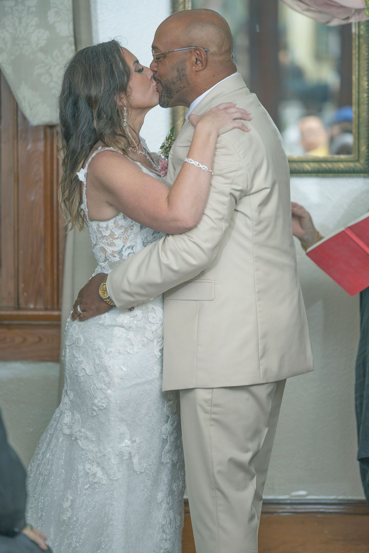 A couple shares their first kiss as newlyweds during their wedding ceremony, with the bride in white lace and groom in tan suit.