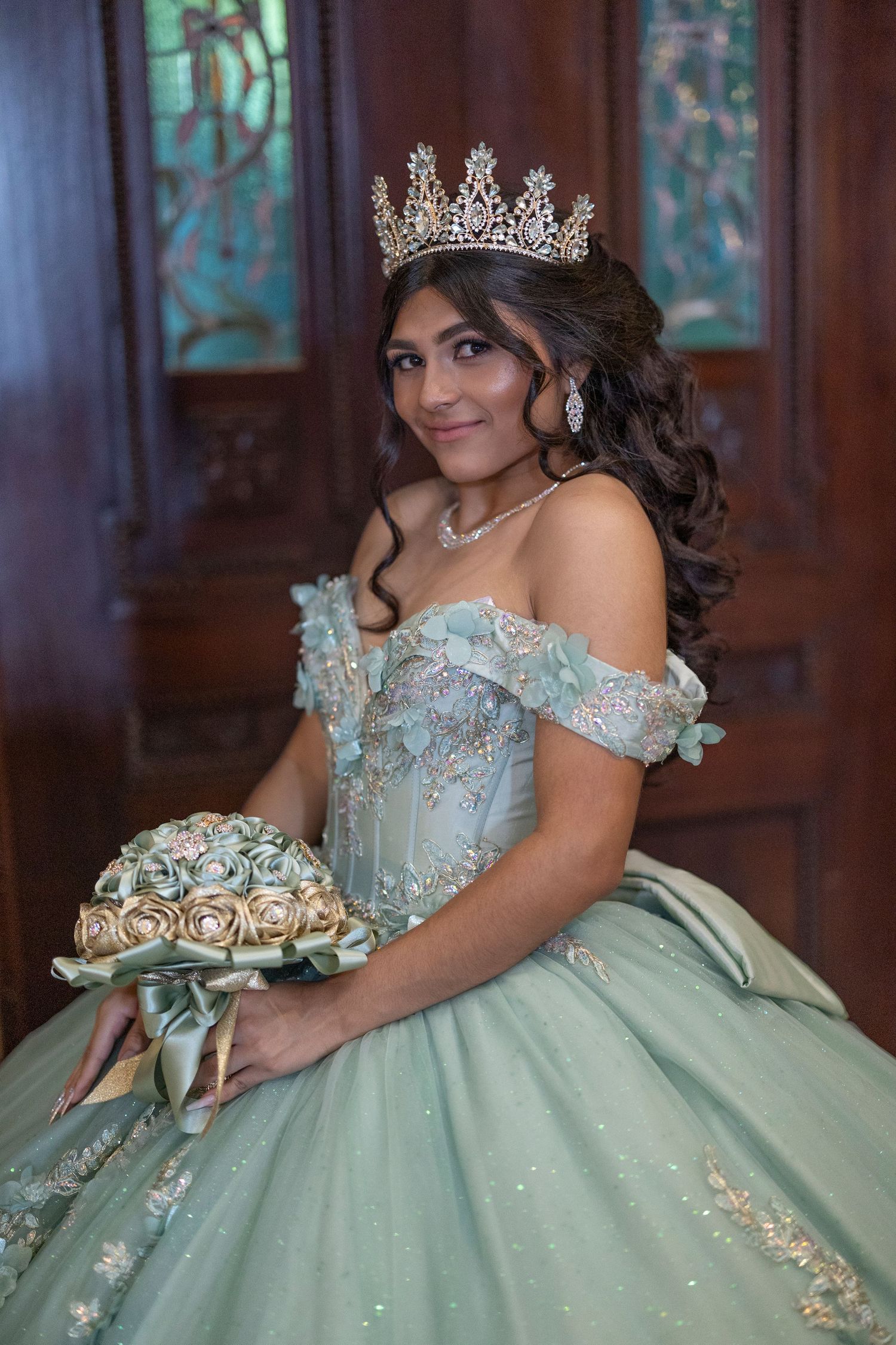A person in a sparkly mint green quinceañera ball gown with off-shoulder design poses elegantly in a formal setting.