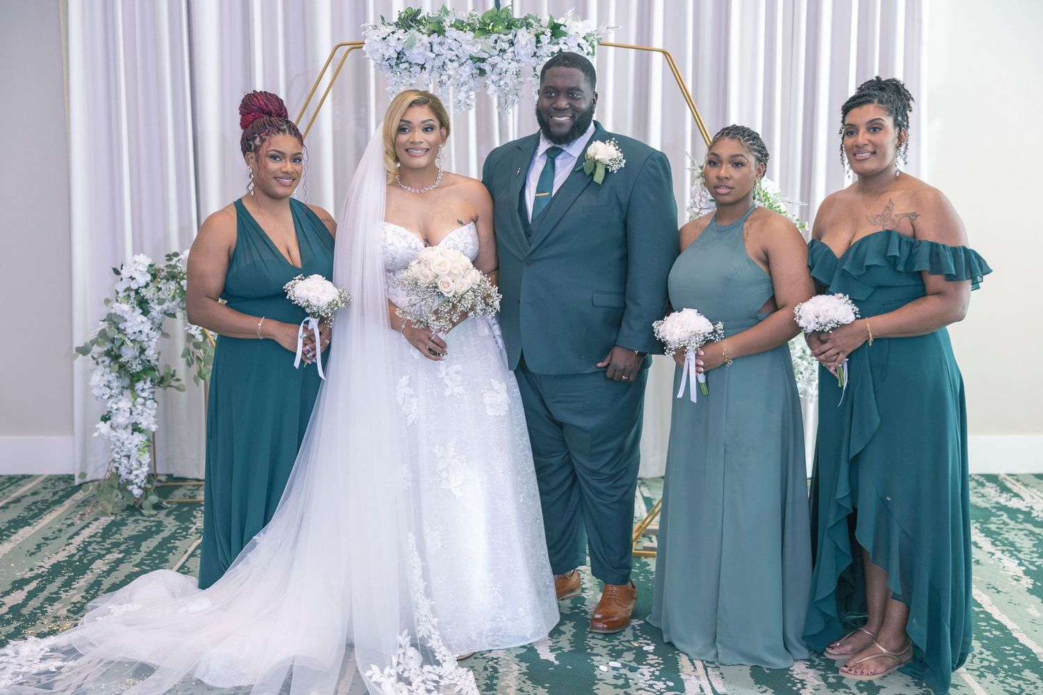 Wedding party in teal dresses and suits pose together with white floral bouquets in an elegant indoor setting.