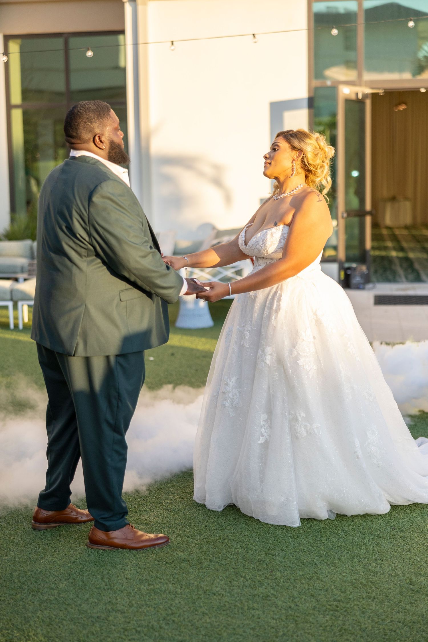 A bride in a sparkly white ballgown and groom in dark suit hold hands during outdoor wedding ceremony on artificial turf.